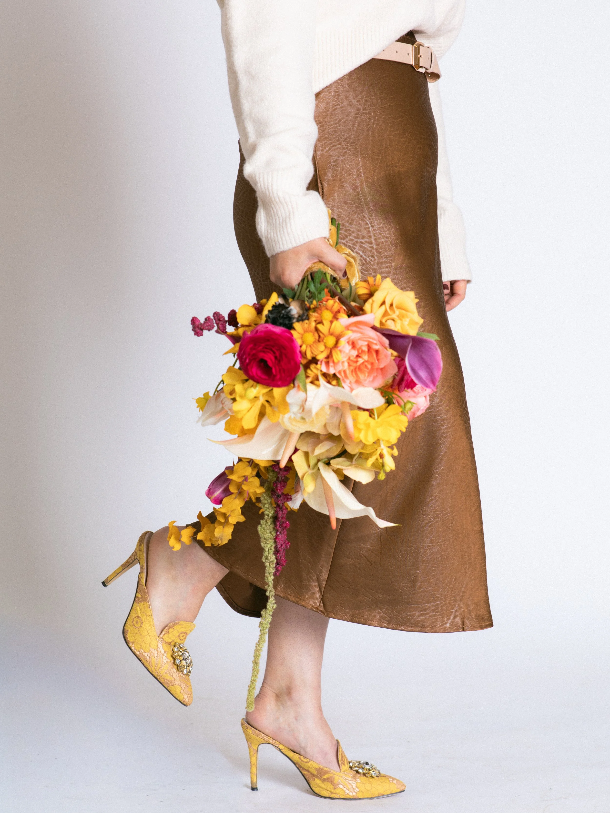 A person wearing yellow lace high heels with embellishments, a brown leather skirt, and a cream sweater, holding a colorful bouquet of flowers.
