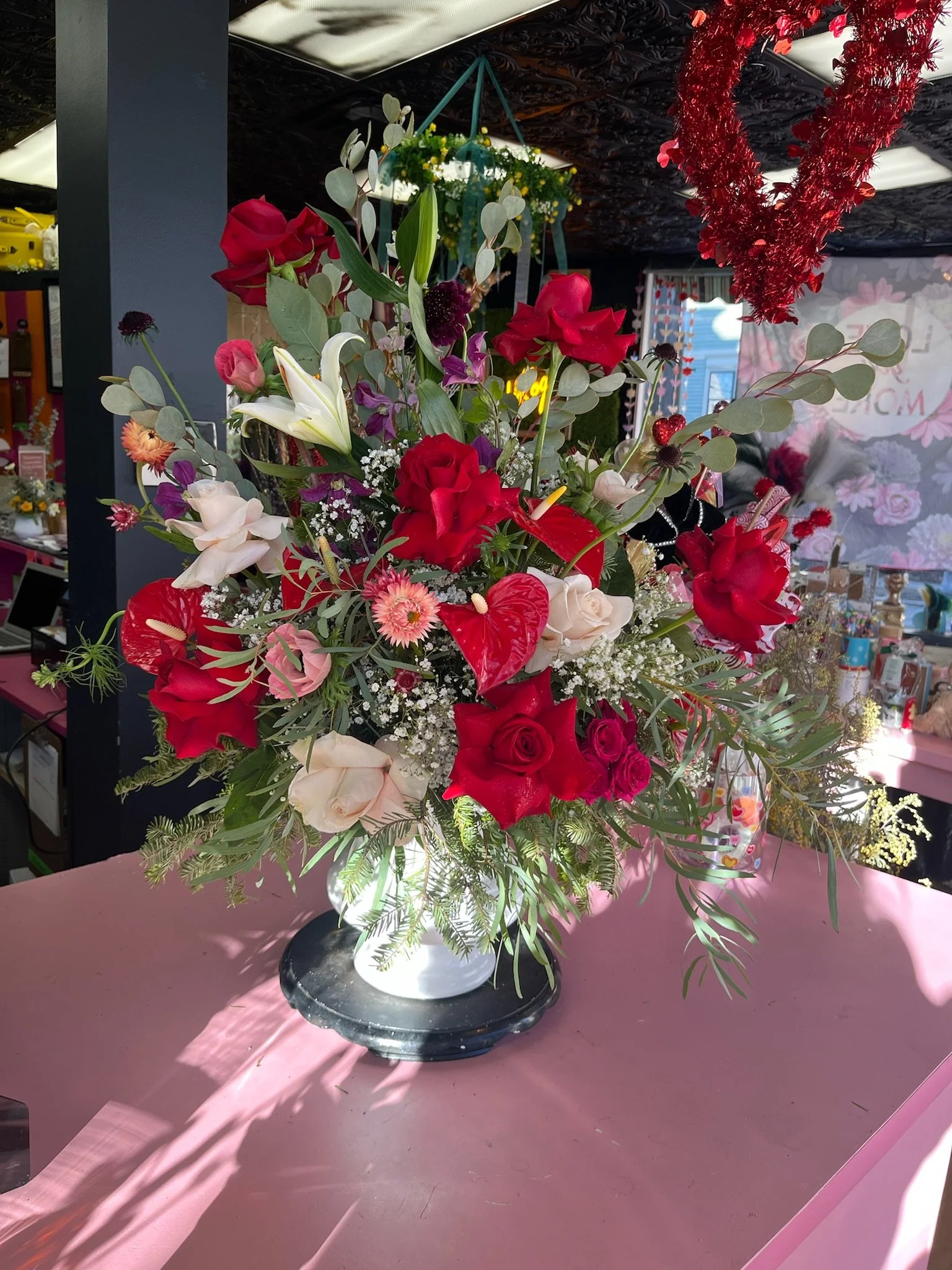 A colorful floral arrangement in a white vase on a pink table, featuring white lilies, red roses, pink and purple flowers, and various green foliage.