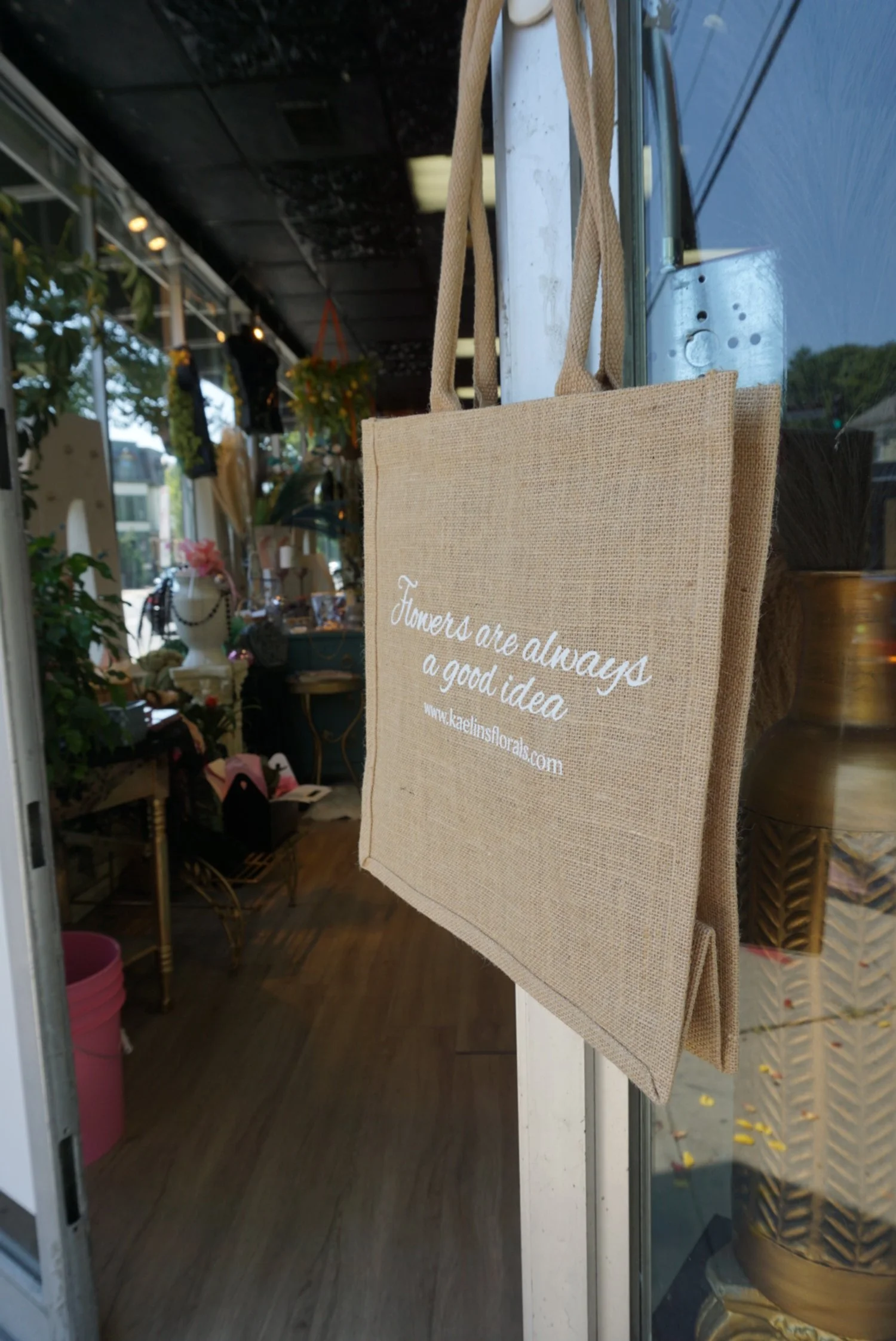 A beige tote bag hanging on a metal hook outside a flower shop. The tote has white text that reads, 'Flowers are always a good idea' and includes a website URL, www.kaelinsflorals.com.