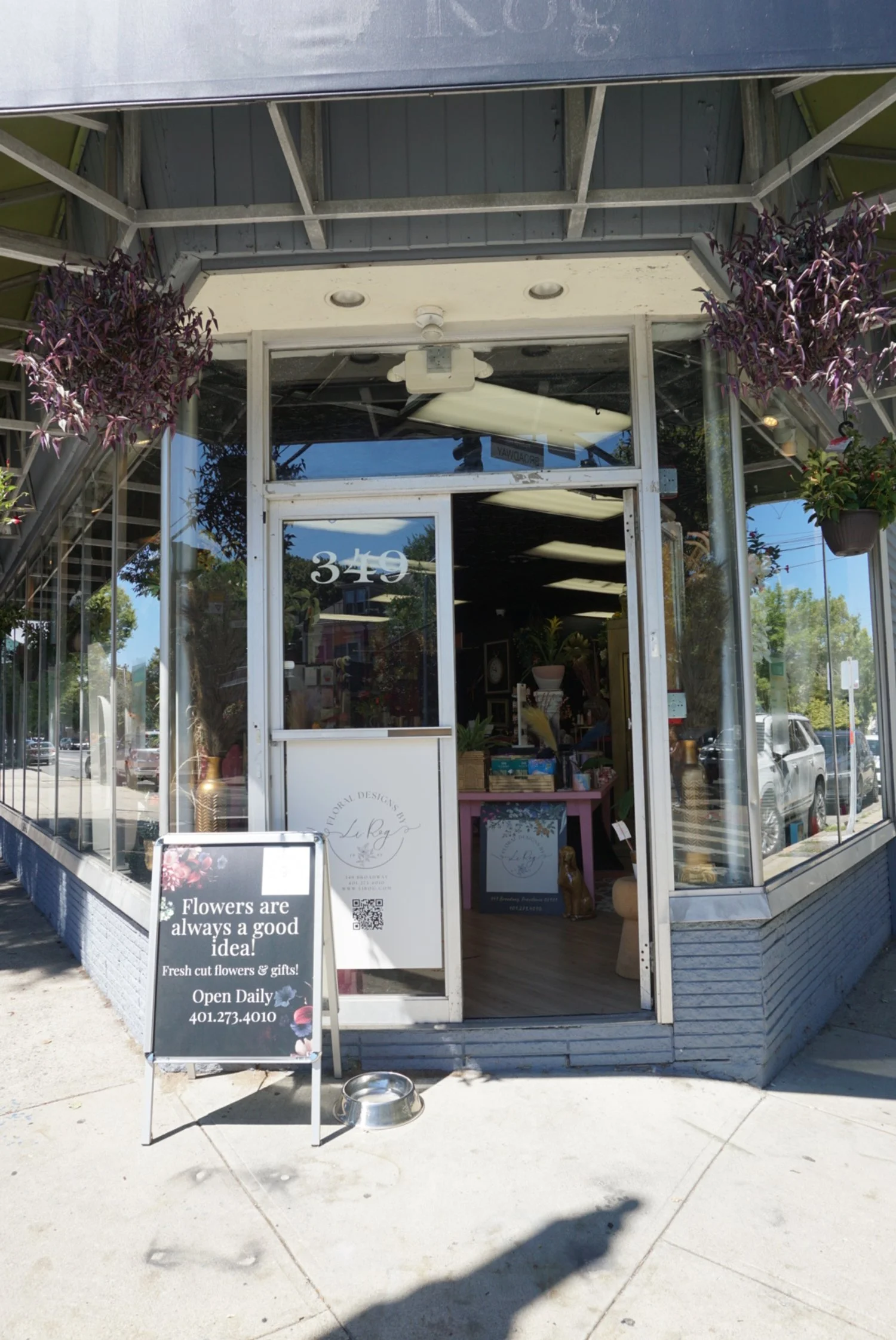 Front entrance of a flower shop with glass windows, displaying potted plants and flowers inside. Signboard outside states, 'Flowers are always a good idea! Fresh cut flowers & gifts! Open Daily 401.273.4010.'