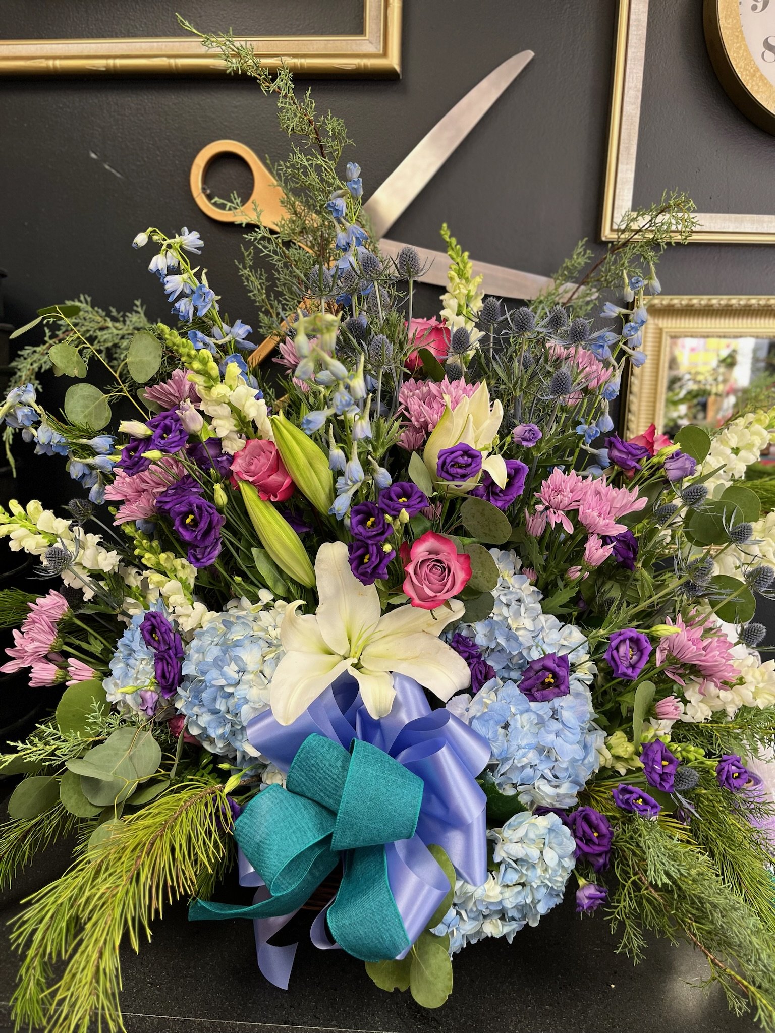 A colorful floral arrangement featuring white lilies, pink roses, purple lisianthus, blue hydrangeas, and various greenery with a blue ribbon bow.