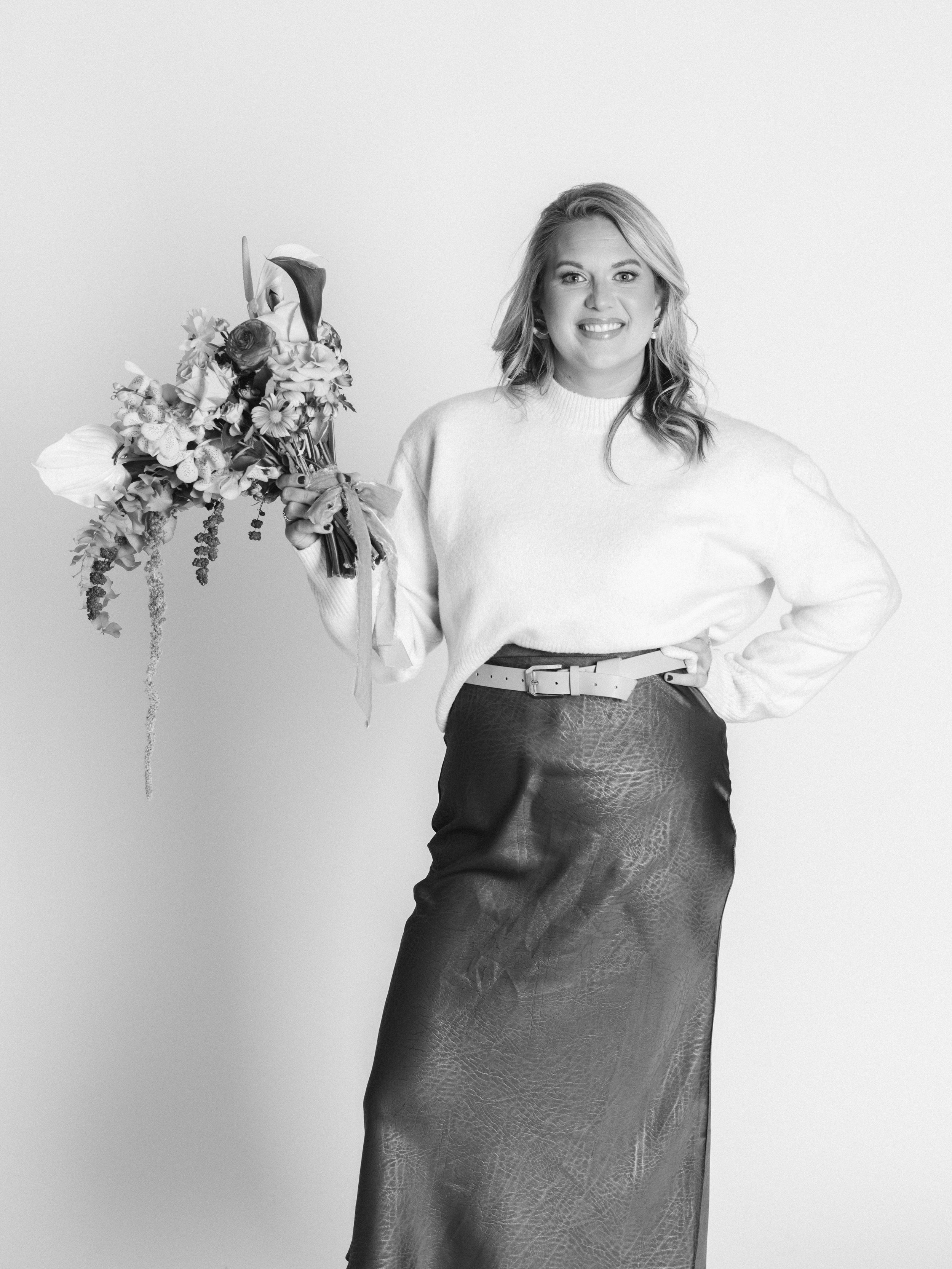 A woman smiling and holding a bouquet of flowers, wearing a light-colored sweater and a long skirt.