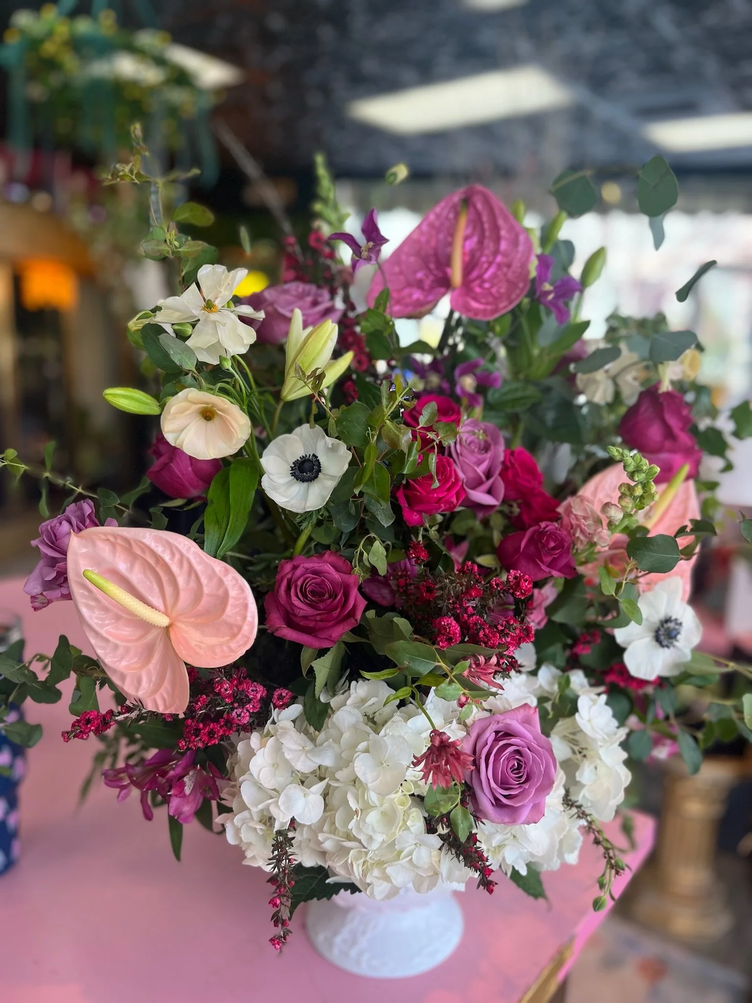 A colorful bouquet of various flowers, including roses, hydrangeas, anthuriums, and anemones, arranged in a white vase on a pink surface.
