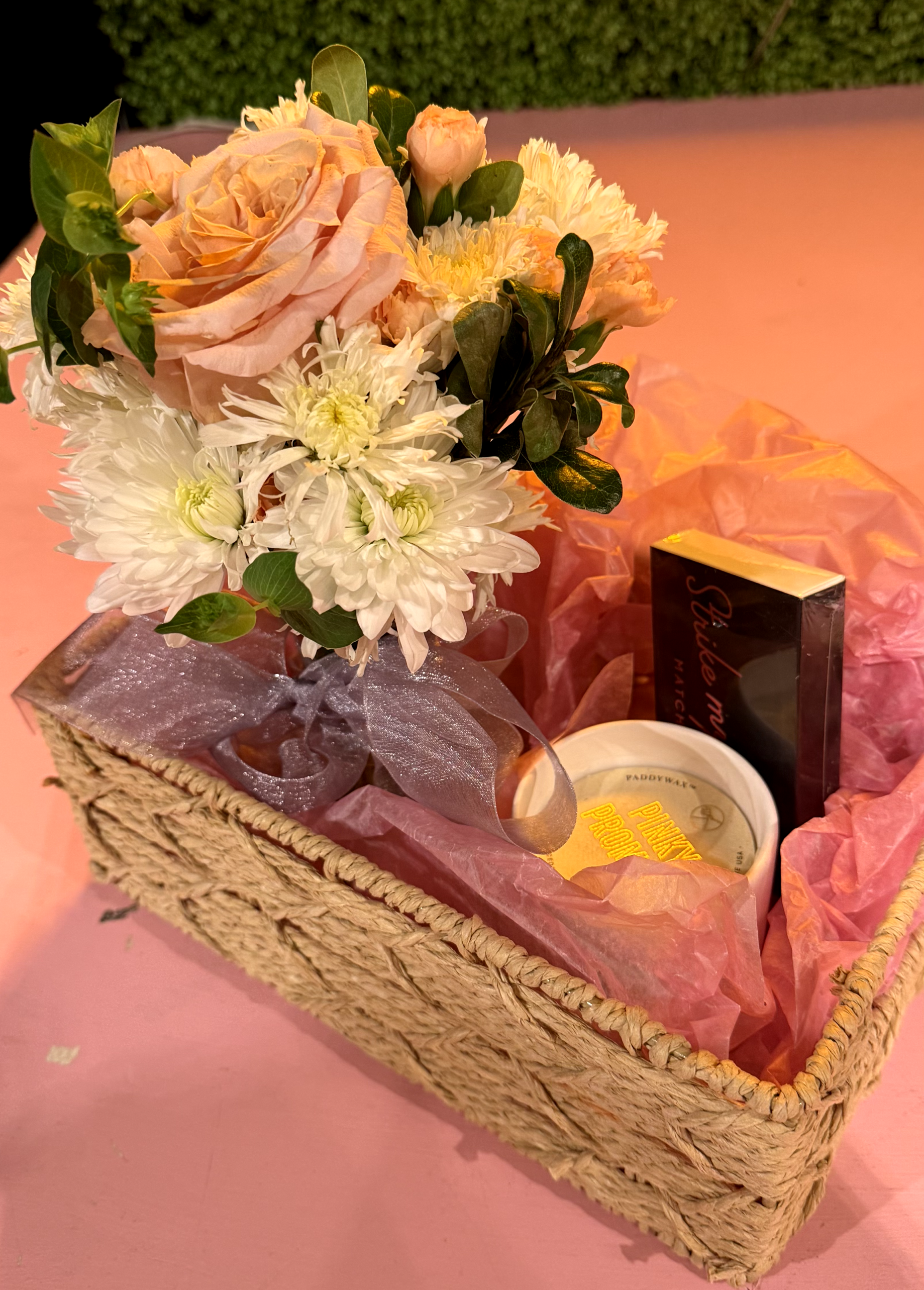 A gift basket with a bouquet of pink roses, white chrysanthemums, and green leaves, lined with pink tissue paper, containing a small box of chocolates, a candle, and a book.