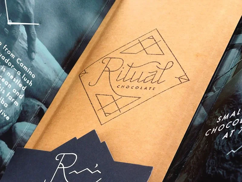A brown paper package with a black logo that reads 'Ritual Chocolate' in cursive, inside a diamond-shaped border with geometric accents.