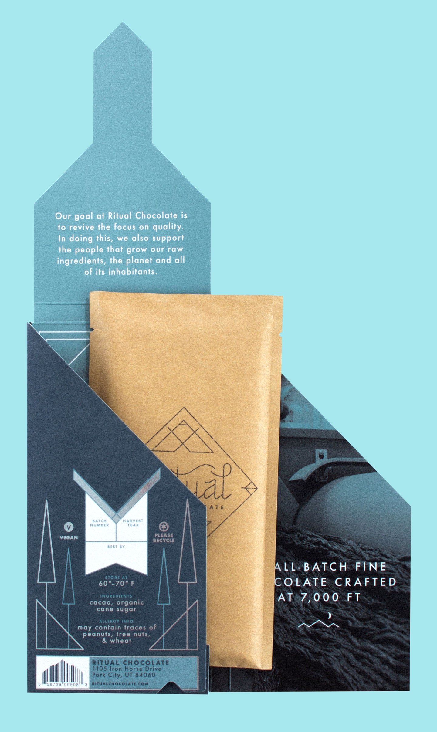 Close-up of a chocolate bar packaging with a mountain silhouette and a tan paper bag on top. The packaging displays information about Vegan, batch number, harvest year, ingredients including cacao, organic cane sugar, and allergy info. The background