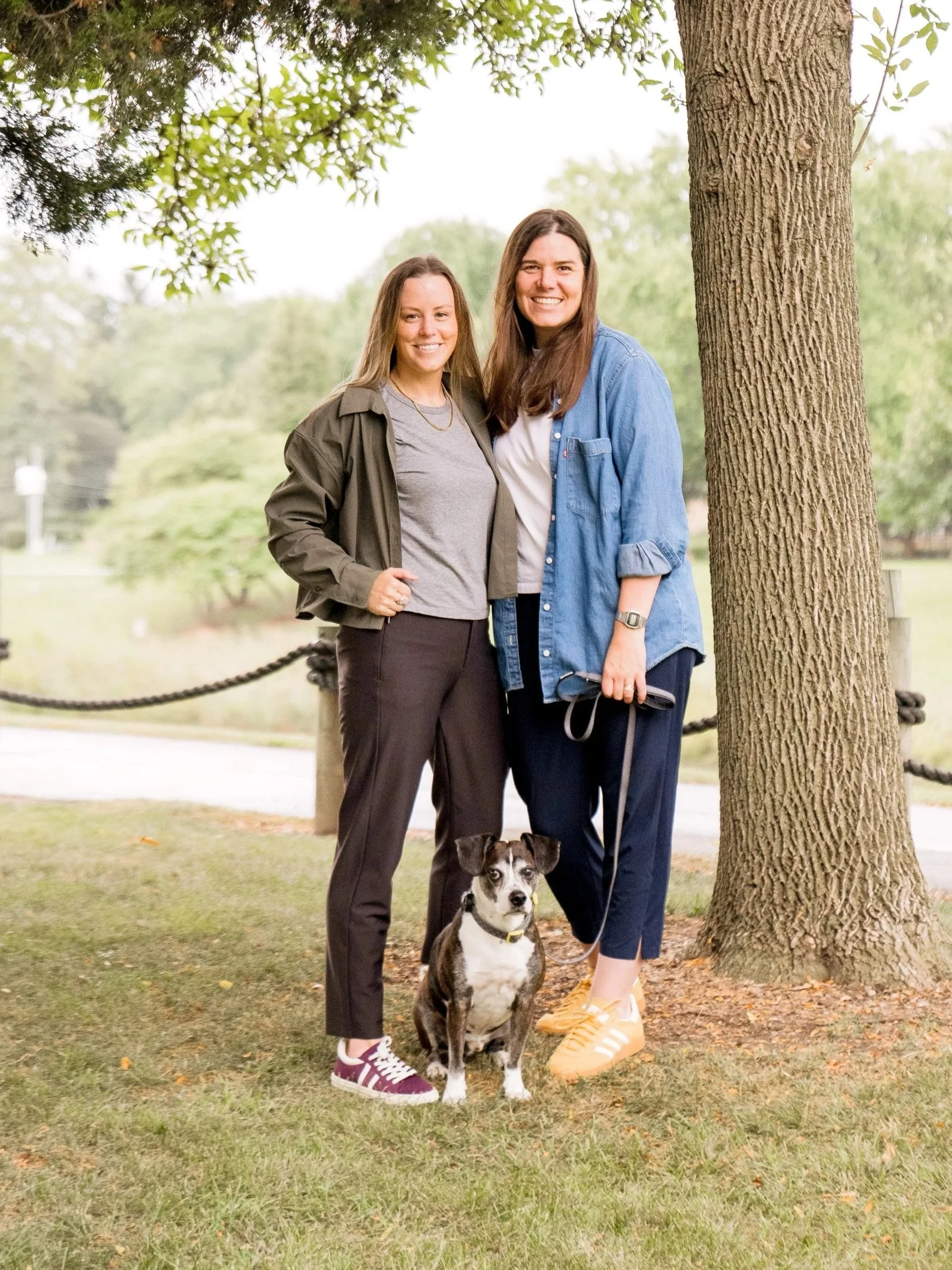 Two smiling women standing outdoors with a dog in front of a tree and greenery.