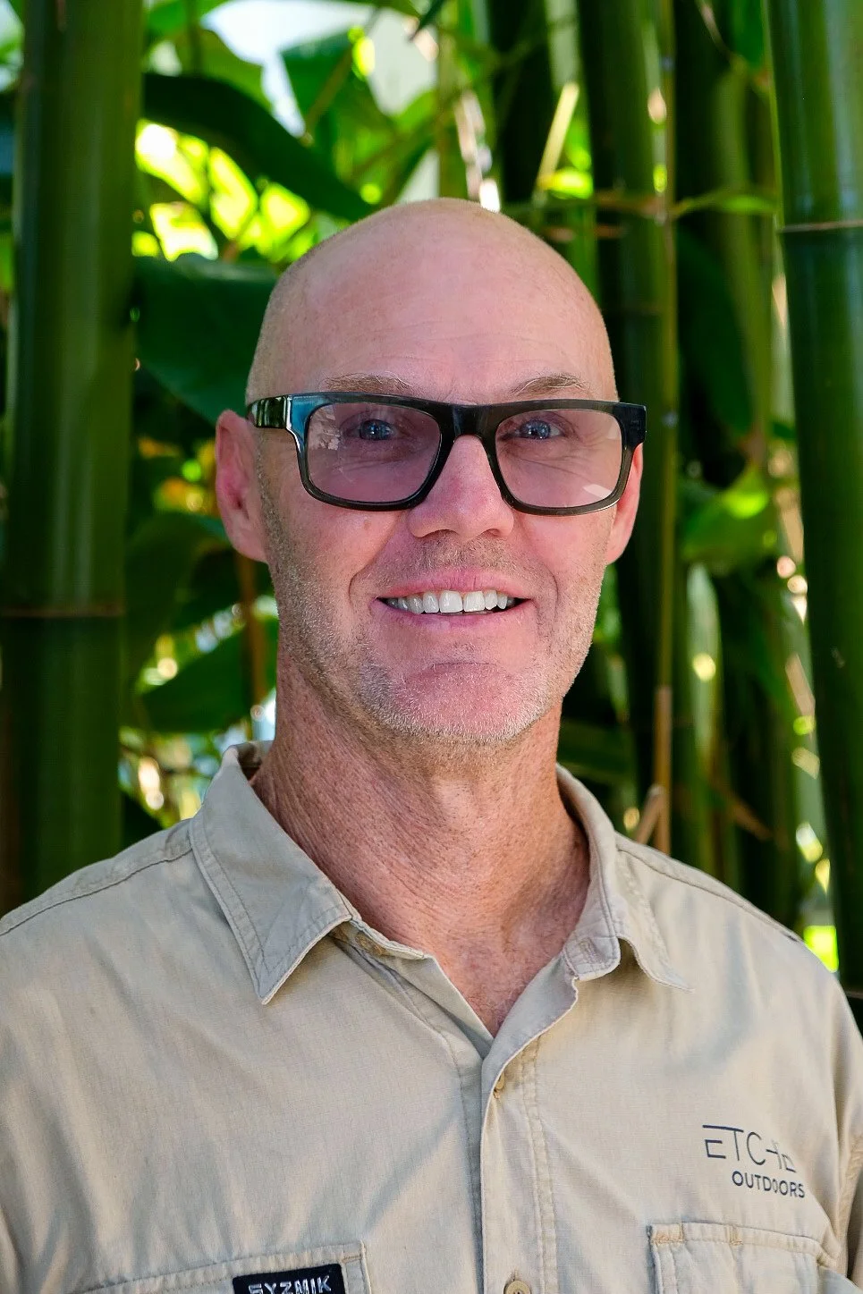 A smiling man wearing glasses and a beige shirt standing outdoors in front of green bamboo plants.