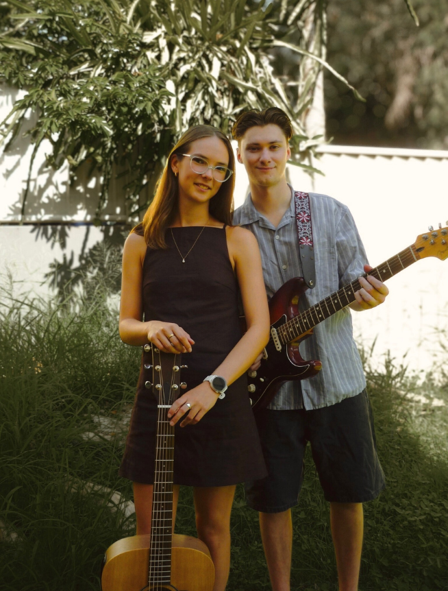 A young woman and young man stand outdoors in a garden with greenery, holding guitars. The woman holds an acoustic guitar, and the man holds an electric guitar. They are smiling at the camera.