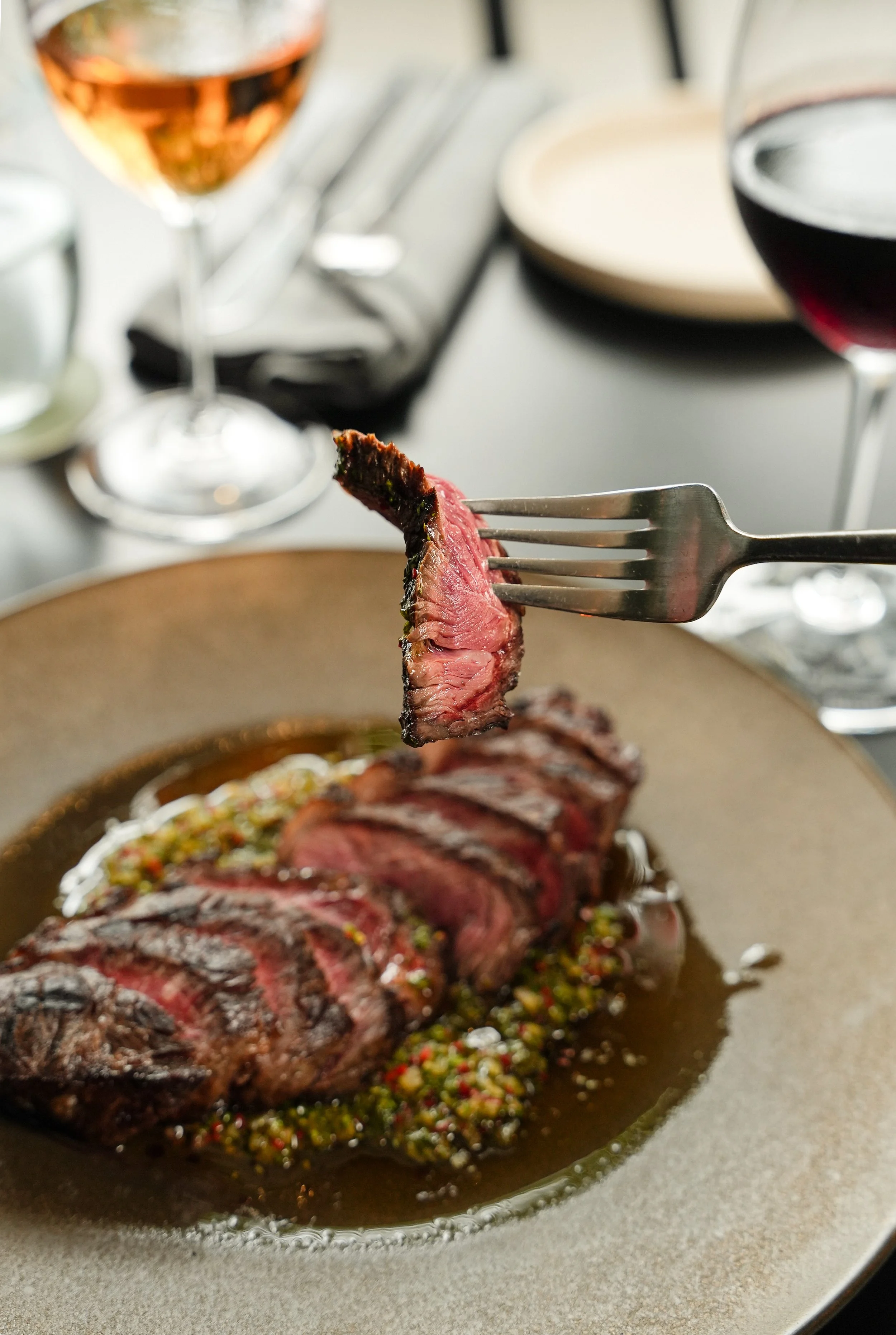 Slices of cooked steak on a plate, with a fork holding a piece, garnished with herbs and served with sauce. Two glasses of wine, one rosé and one red, are in the background.