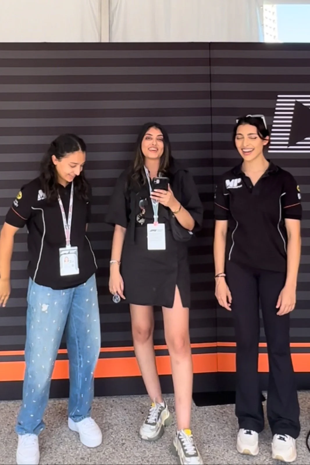 Three women standing together against a black and gray striped wall, all wearing black shirts with logos, smiling and laughing, one holding a smartphone.