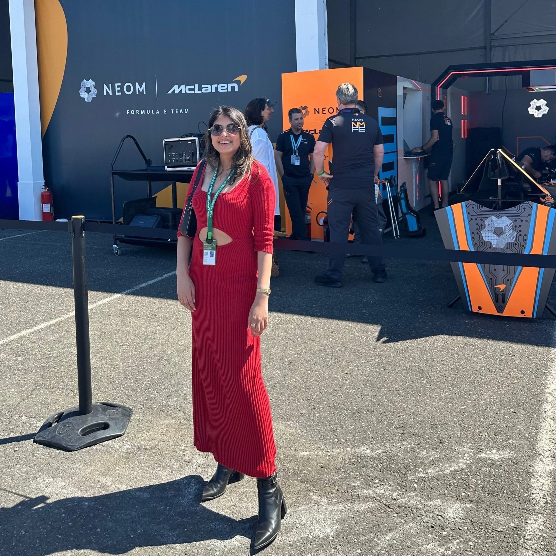 A woman in a red dress and black boots smiling and standing outside in front of a Formula 1 race team garage. The garage has signs for NEOM and McLaren, with people working inside and around the area.