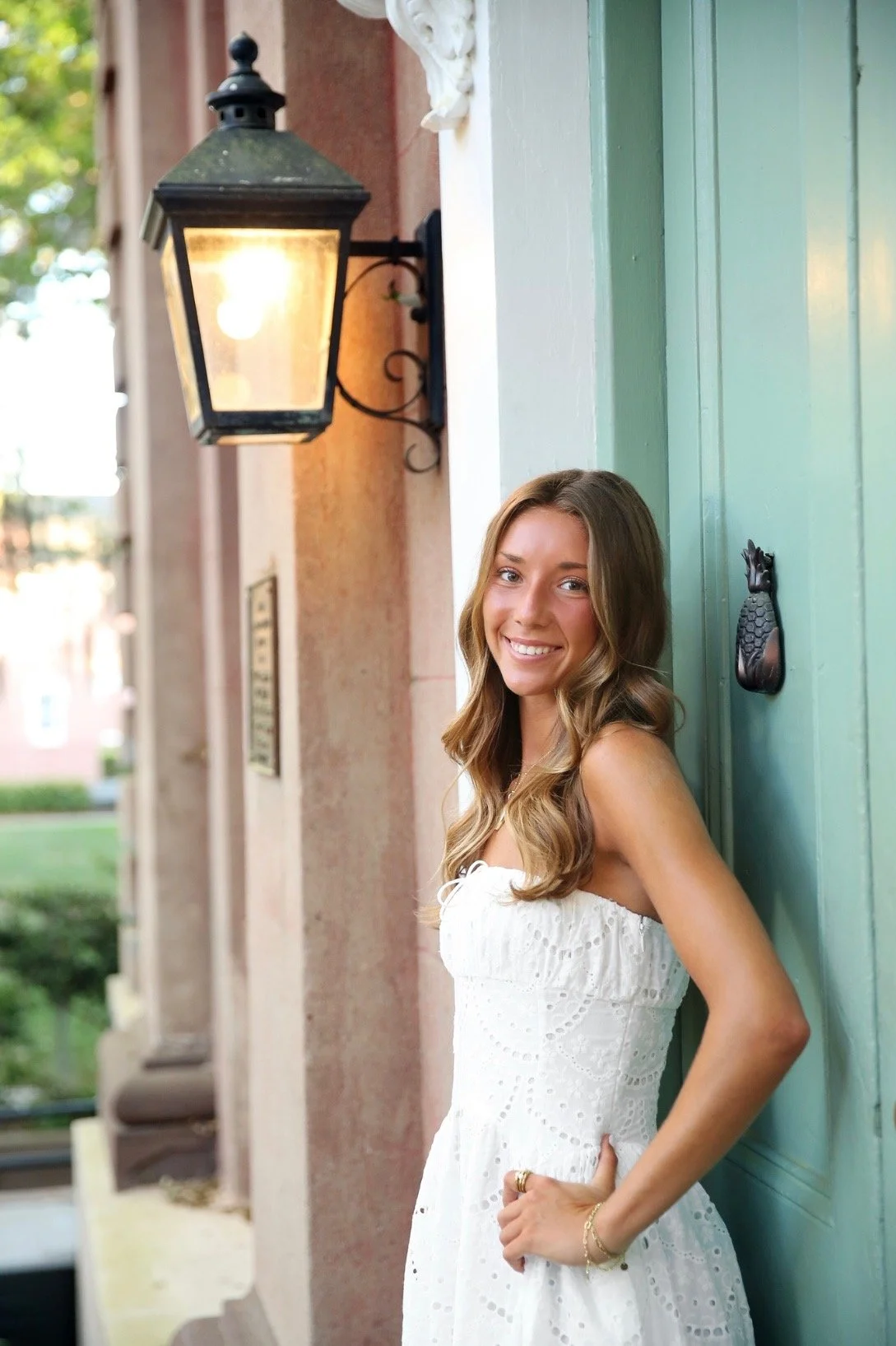 A young woman with long, wavy brown hair smiling and leaning against a green door, wearing a white sleeveless dress, outside near a building with a wall-mounted lantern.