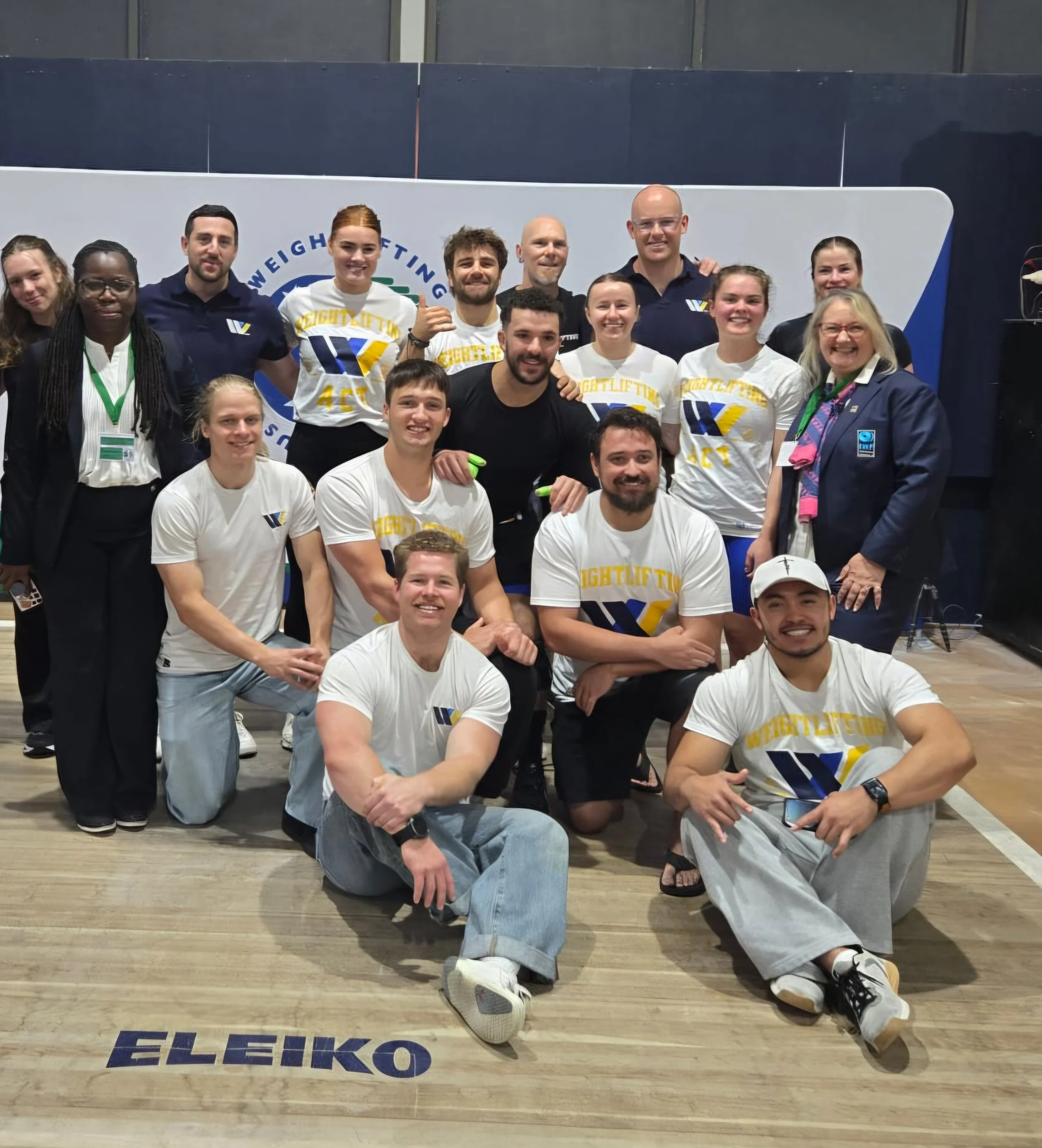Group of people, some wearing sports attire with 'Weightlifting' and 'ACT' on shirts, posing together in an indoor sports facility.