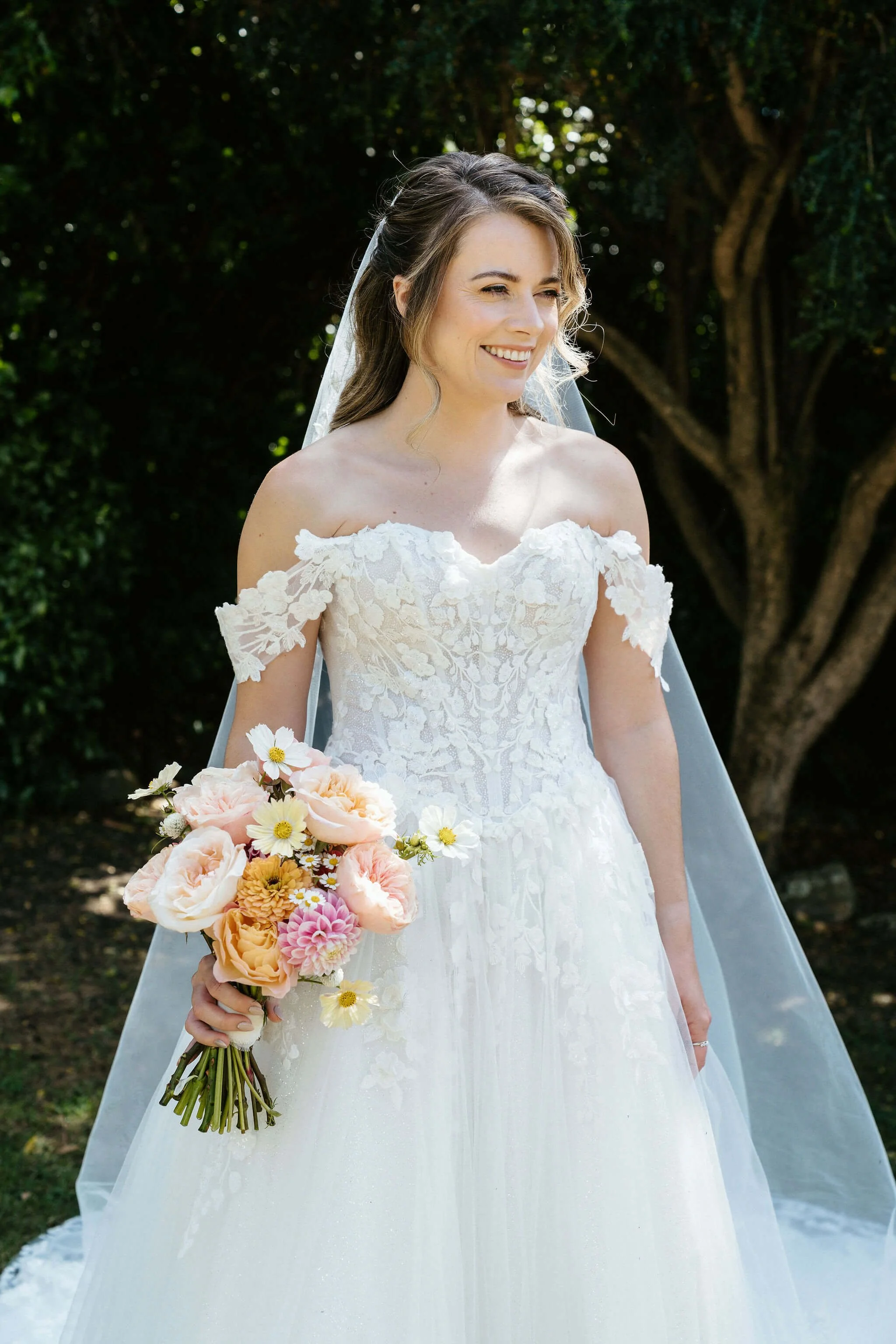 A smiling bride in a white lace wedding gown with off-shoulder floral sleeves, holding a bouquet of pink, peach, and cream flowers, standing outdoors with trees in the background.