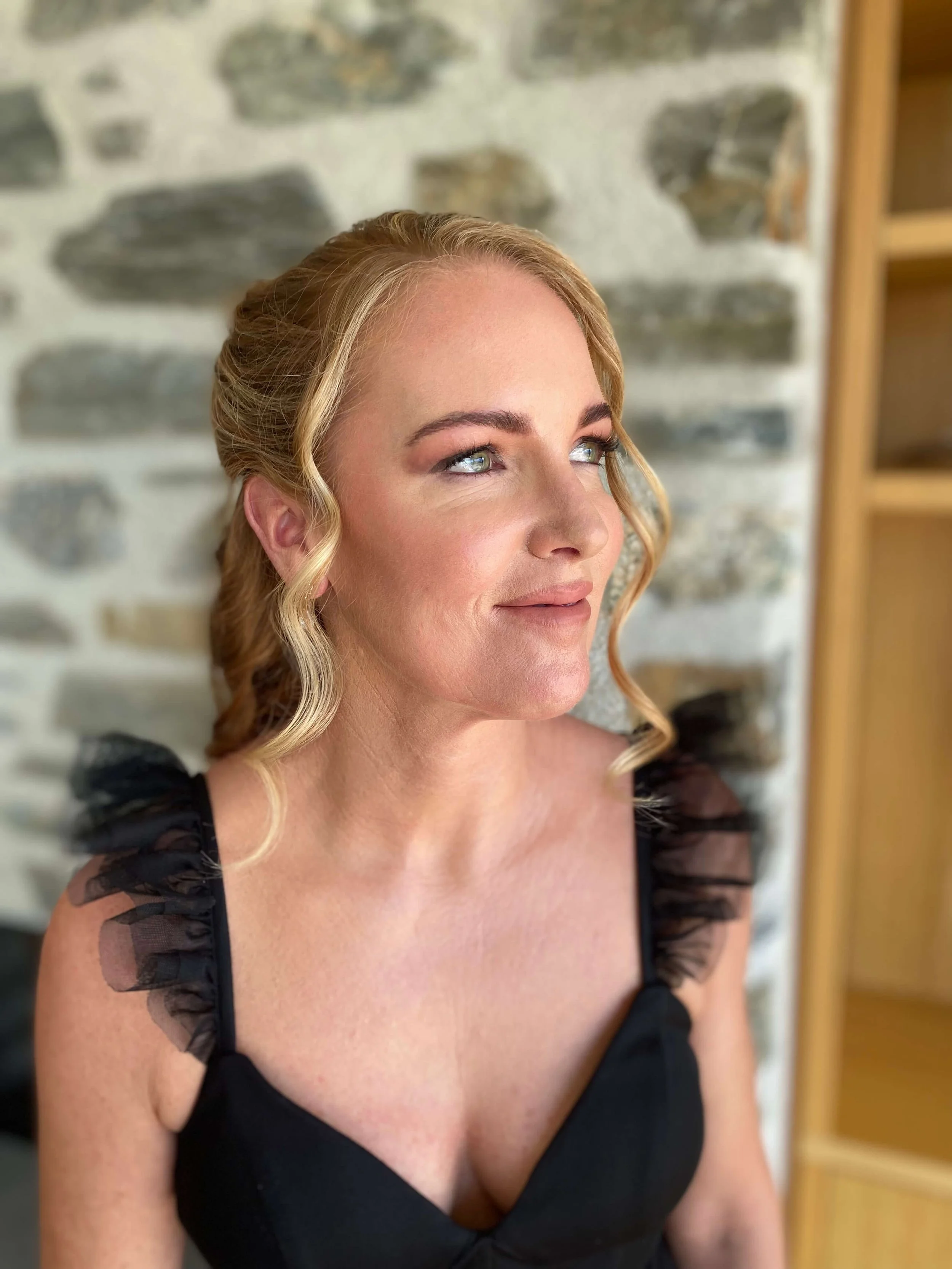 A woman with wavy blonde hair and blue eyes wearing a black dress with ruffled tulle sleeves, standing in front of a stone wall.