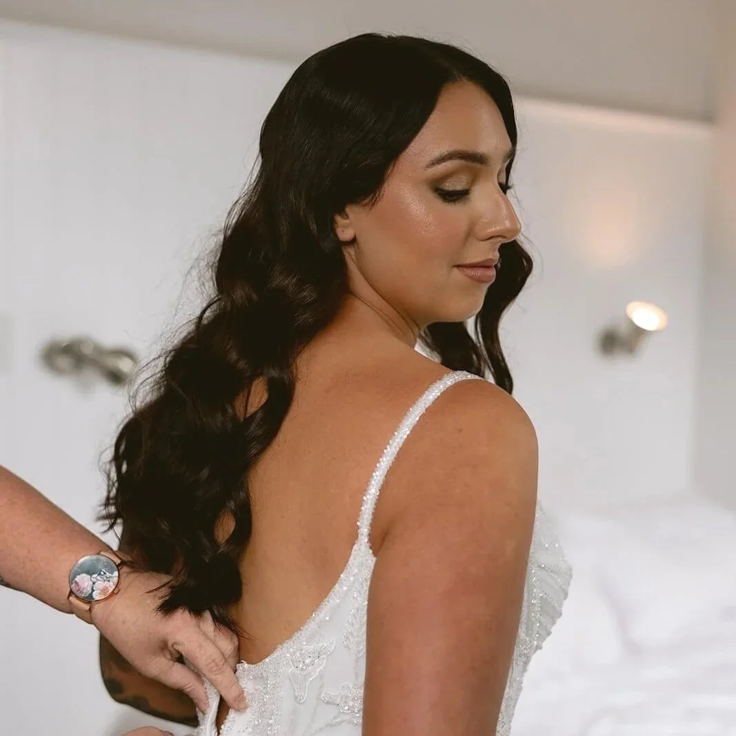 A woman with long, wavy dark hair and tan skin, wearing a white dress with thin straps, is seated as someone adjusts it from behind. She has makeup with neutral tones and soft waves in her hair.