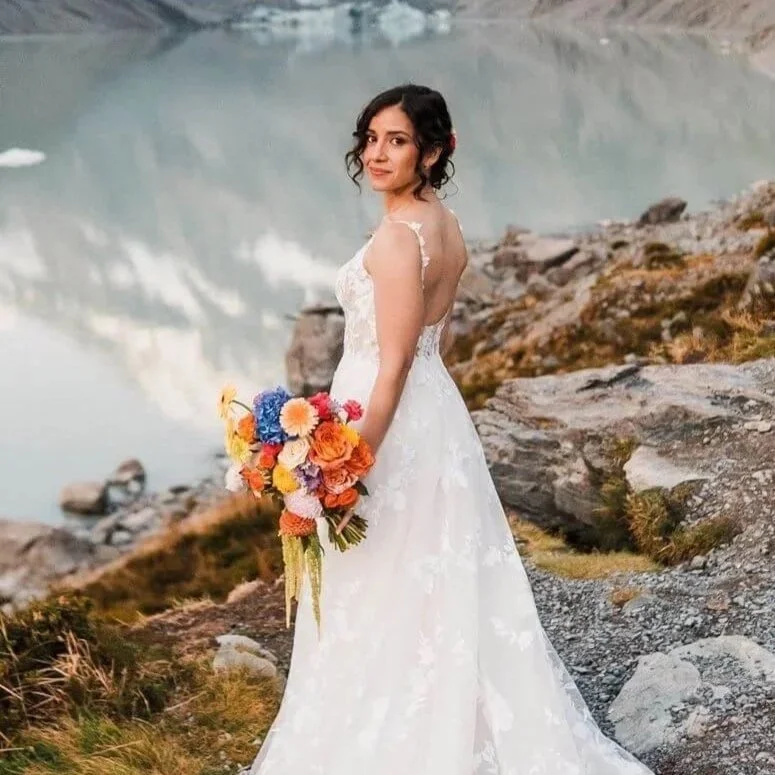A woman in a white wedding dress holding a colorful bouquet of flowers, standing near a body of water with rocky terrain around her.