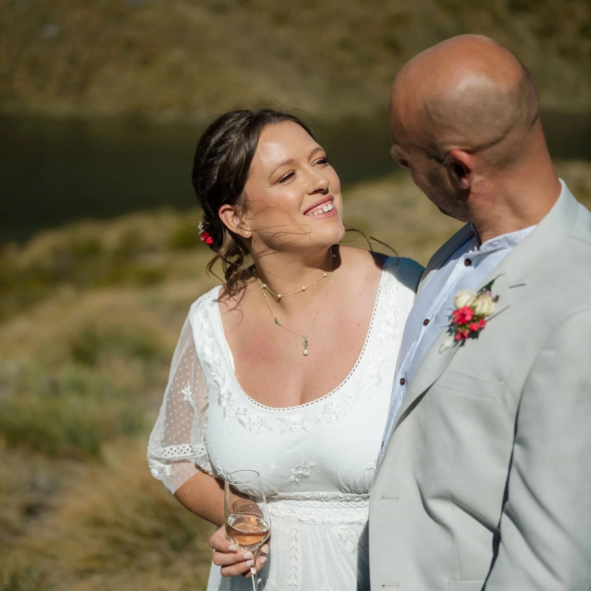 A woman in a white wedding dress smiling at a man in a light-colored suit, holding a glass of champagne outdoors near a body of water.