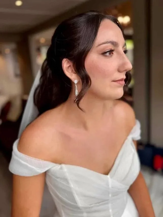 A bride with dark brown hair and earrings, wearing a white off-shoulder wedding gown, standing indoors in a warmly lit room.