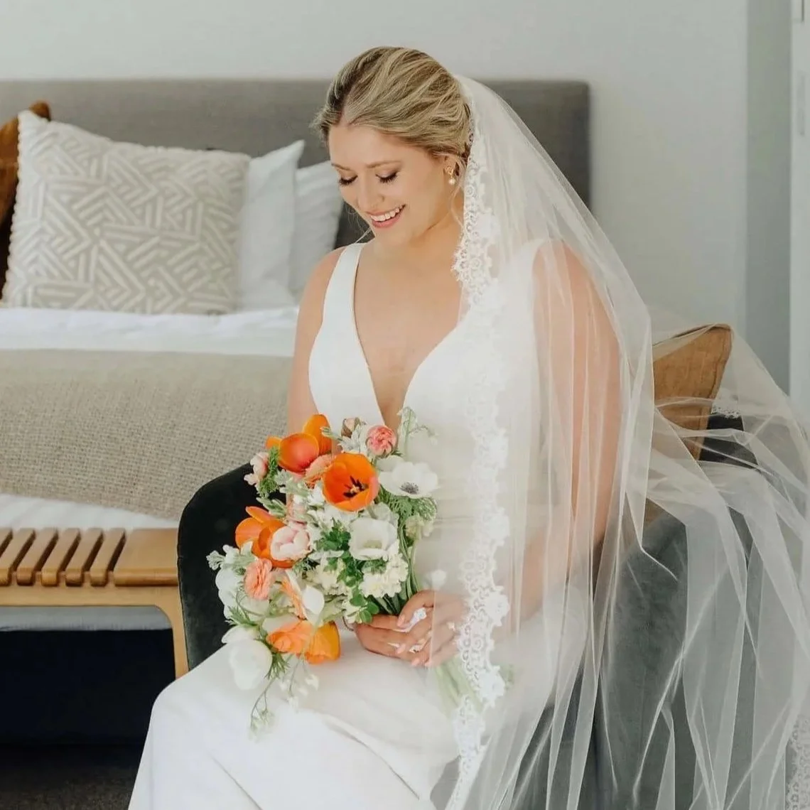 A bride sitting in a modern, minimalistic room holding a bouquet of flowers, wearing a white wedding dress and veil, smiling gently.