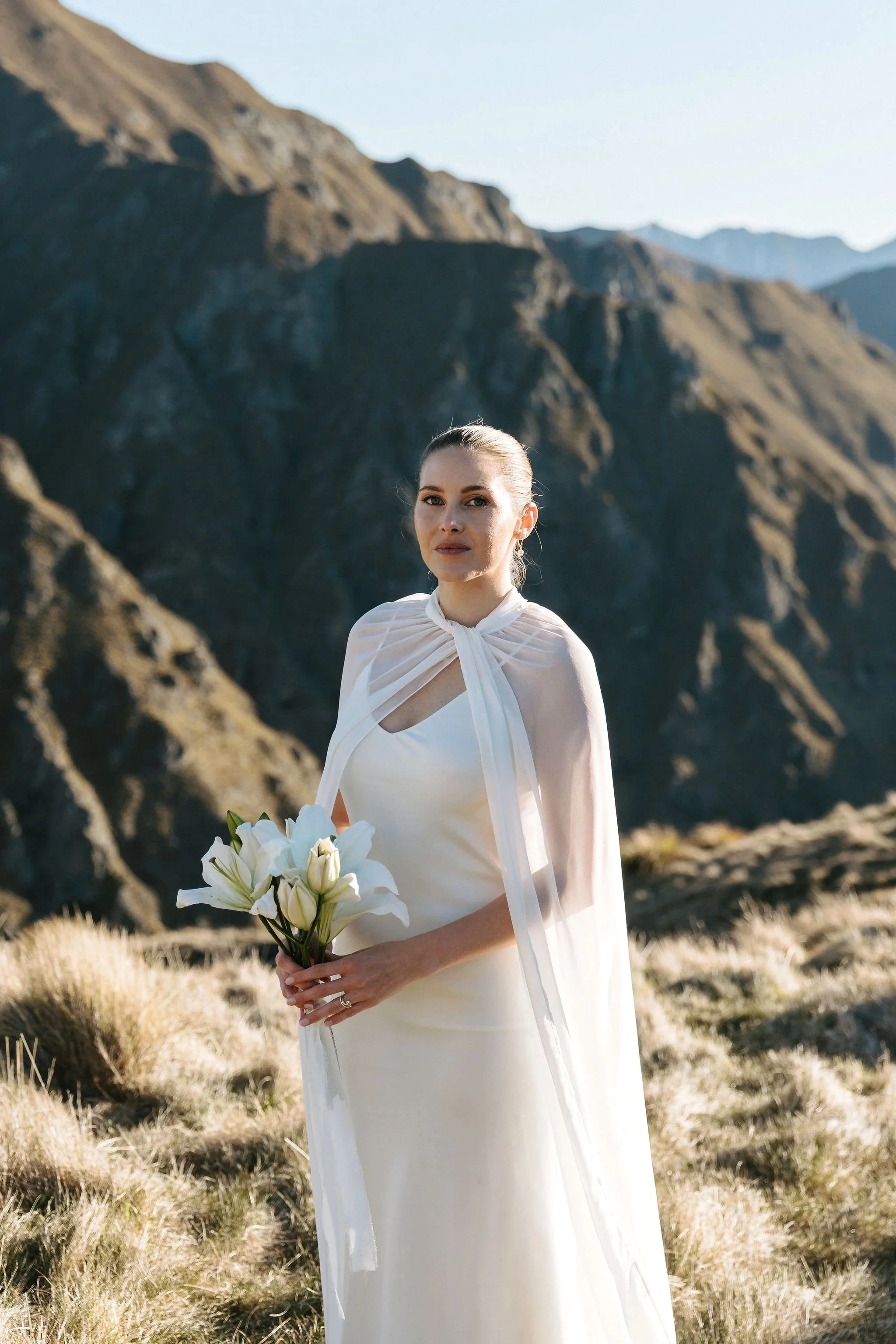 A woman wearing a white dress with a sheer neckline stands outdoors in a mountainous landscape, holding a bouquet of white lilies, with mountains in the background.