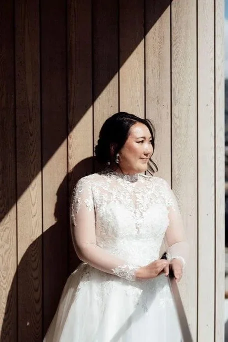 A woman in a white lace wedding dress standing against a wooden wall, with sunlight casting a shadow.