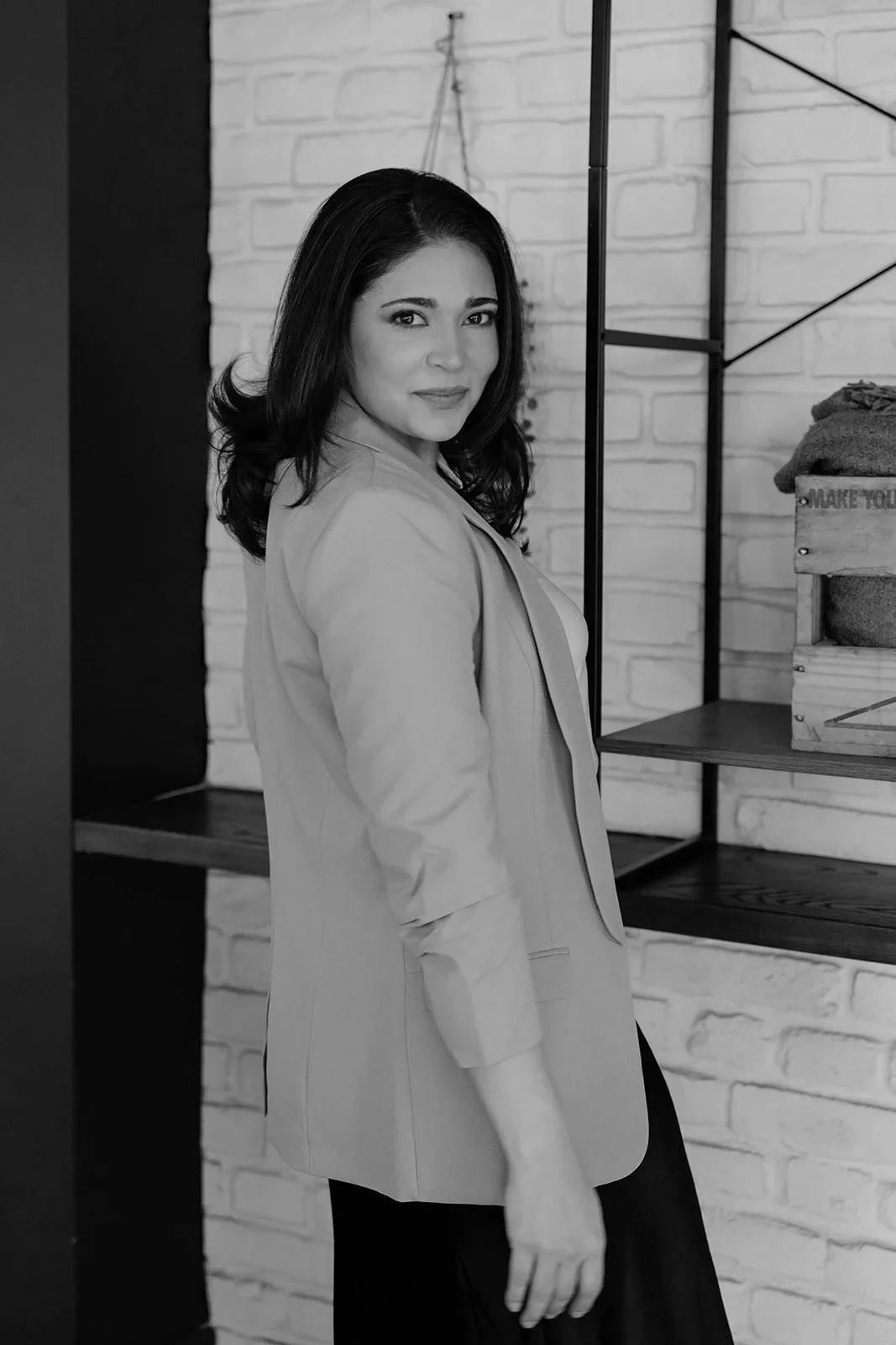 A woman with dark, shoulder-length hair and a light-colored blazer stands near a brick wall and shelving, looking at the camera.