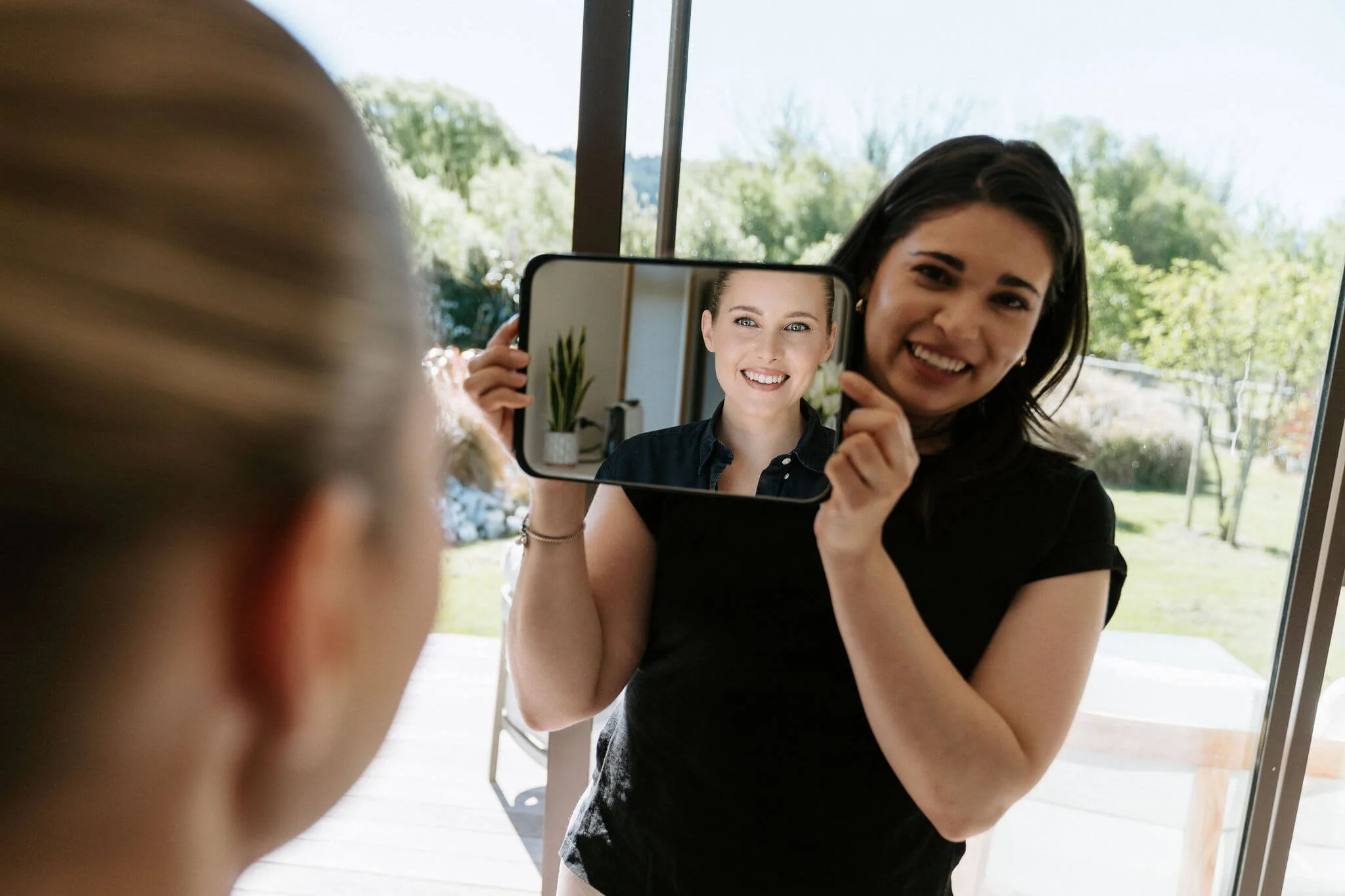 Two women take a photo of each other through a mirror on a glass door, with outdoor scenery visible in the background.