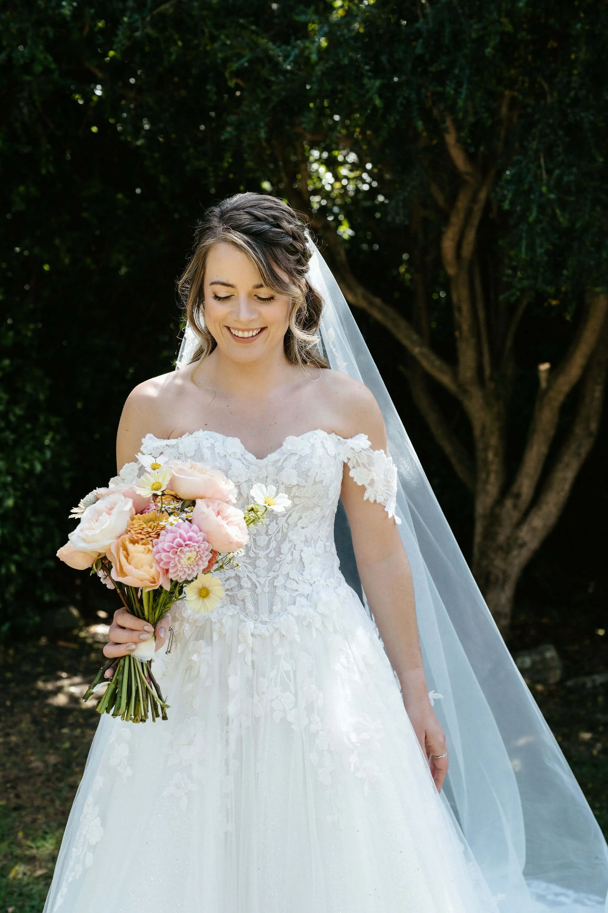 A bride wearing a white lace wedding dress and a veil, holding a bouquet of pink, peach, yellow, and white flowers, standing outdoors in front of a tree with green foliage, smiling with her eyes closed.