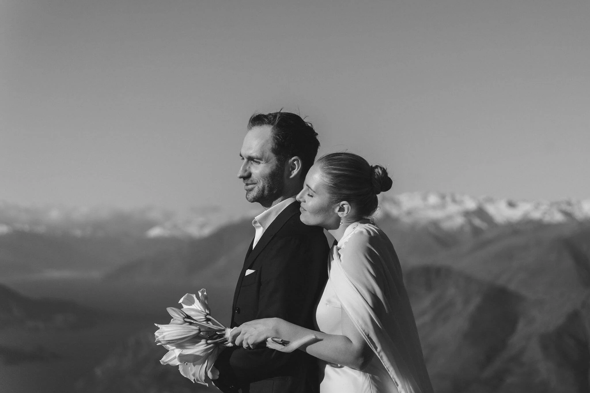 Black and white photograph of a man and woman standing outdoors on a mountain landscape. The man is in a suit holding a bouquet of flowers, and the woman, wearing a light-colored dress and a shawl, is leaning her head on his shoulder.