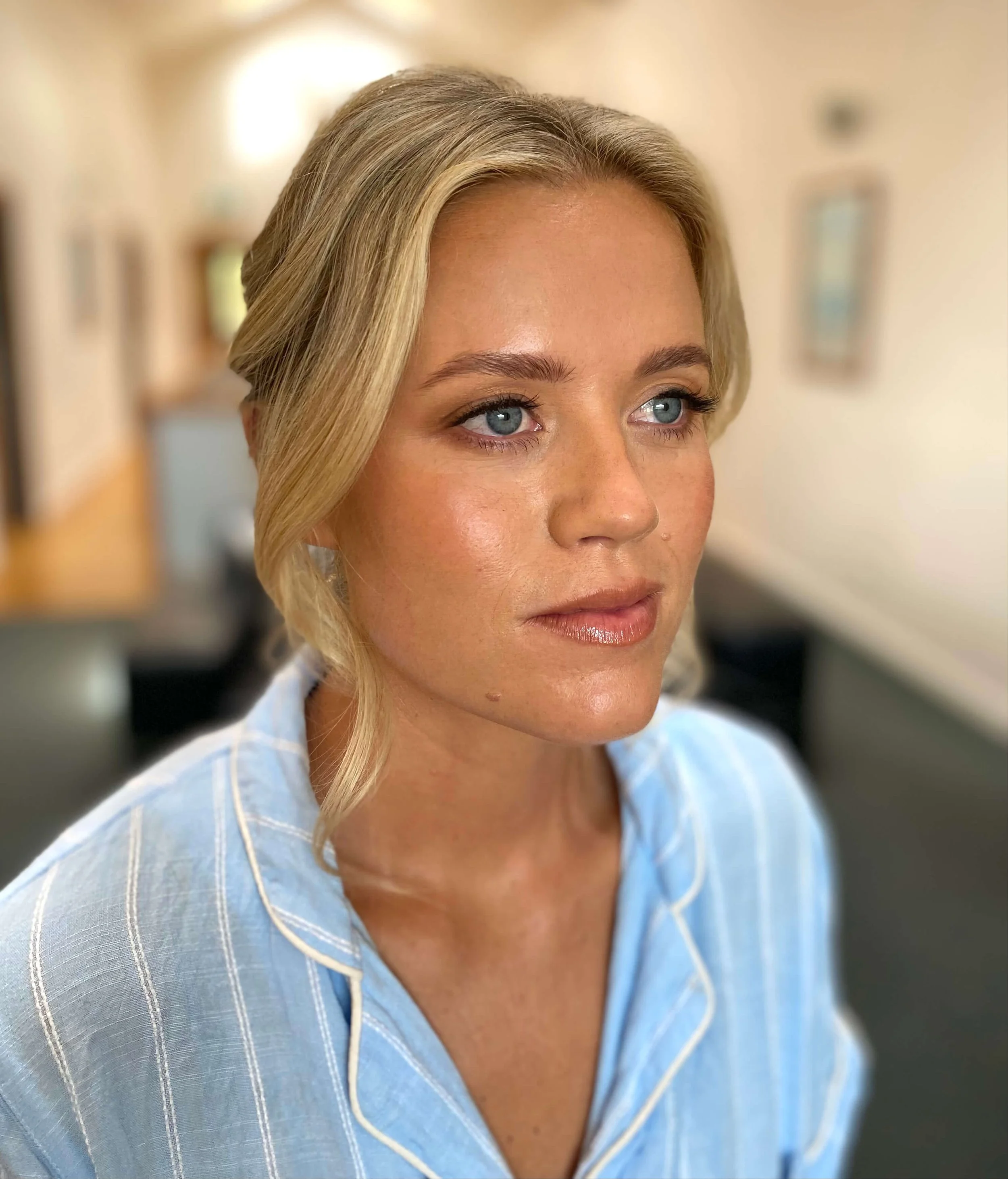 Close-up of a woman with blonde hair, blue eyes, wearing a blue and white striped shirt, in an indoor setting with blurred background.