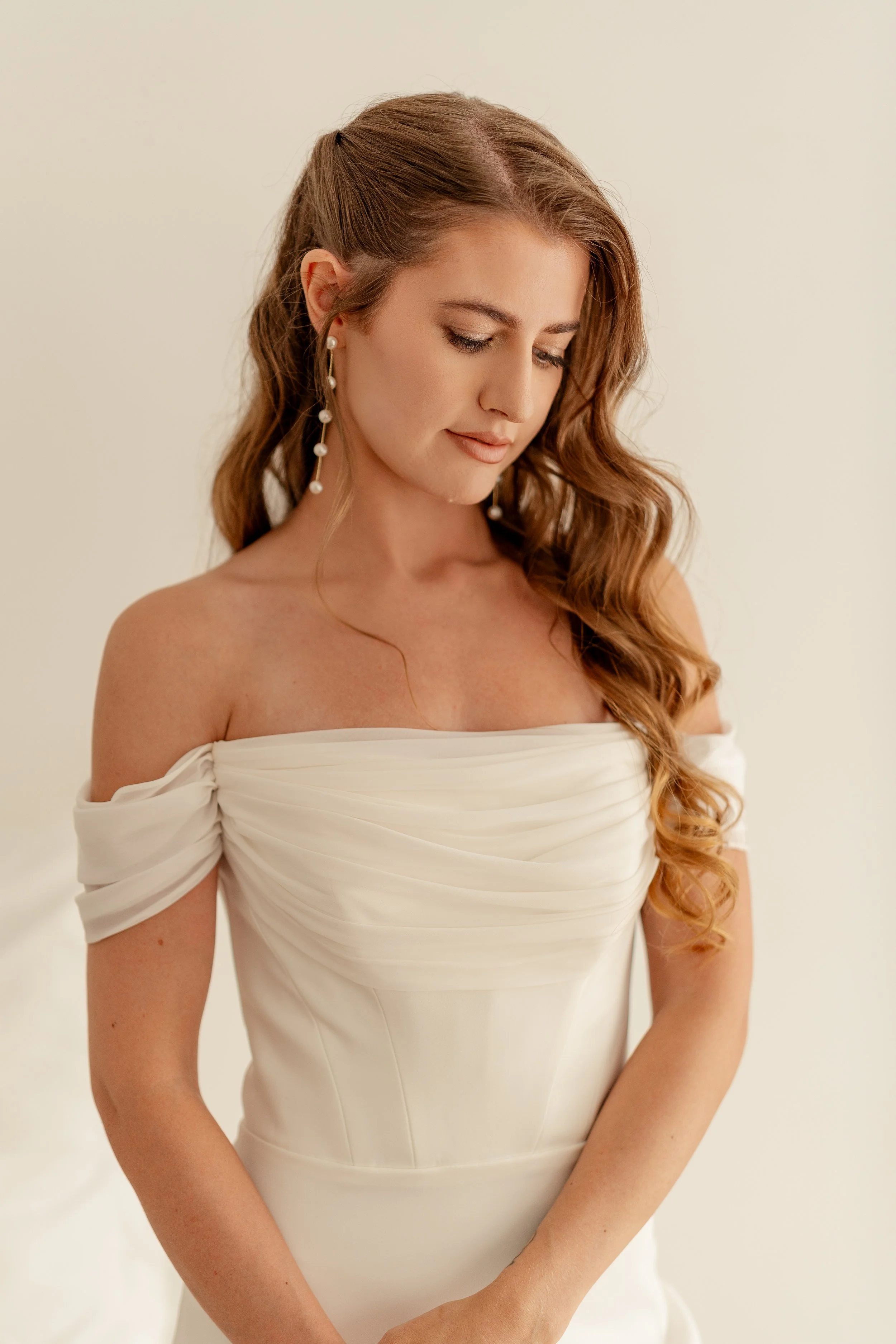 A woman with long wavy auburn hair wearing pearl earrings and an off-the-shoulder white dress, looking down with a gentle smile, against a plain light background.