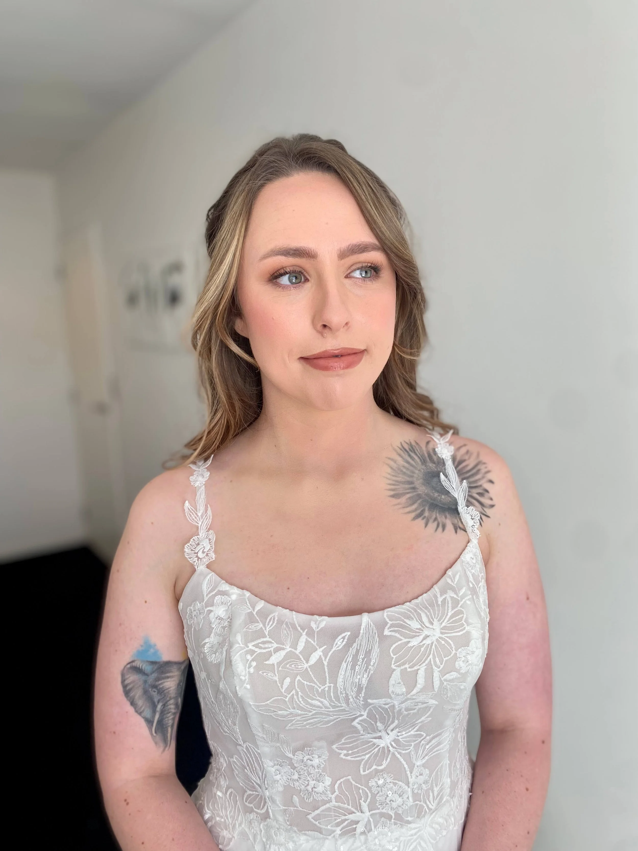 A young woman with wavy brown hair, blue eyes, and fair skin, wearing a white dress with floral embroidery, standing indoors against a plain wall.