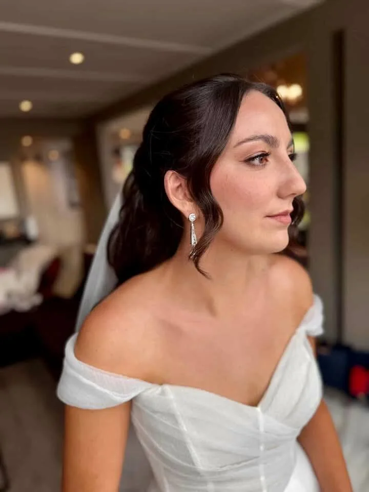 A bride with dark brown hair and earrings, wearing a white off-shoulder wedding gown, standing indoors in a warmly lit room.