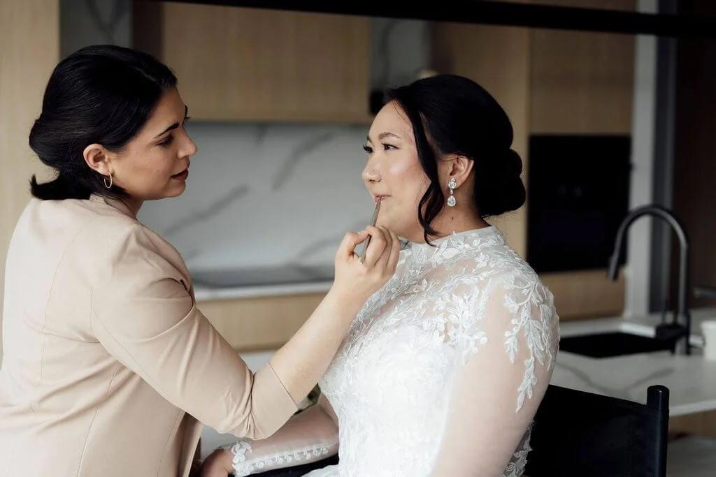 A woman applying makeup to a bride dressed in a lace wedding gown, with a kitchen in the background.