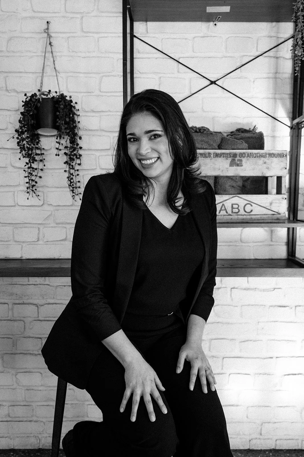A smiling woman in black attire sitting on a chair in front of a white brick wall with hanging plant and shelves.
