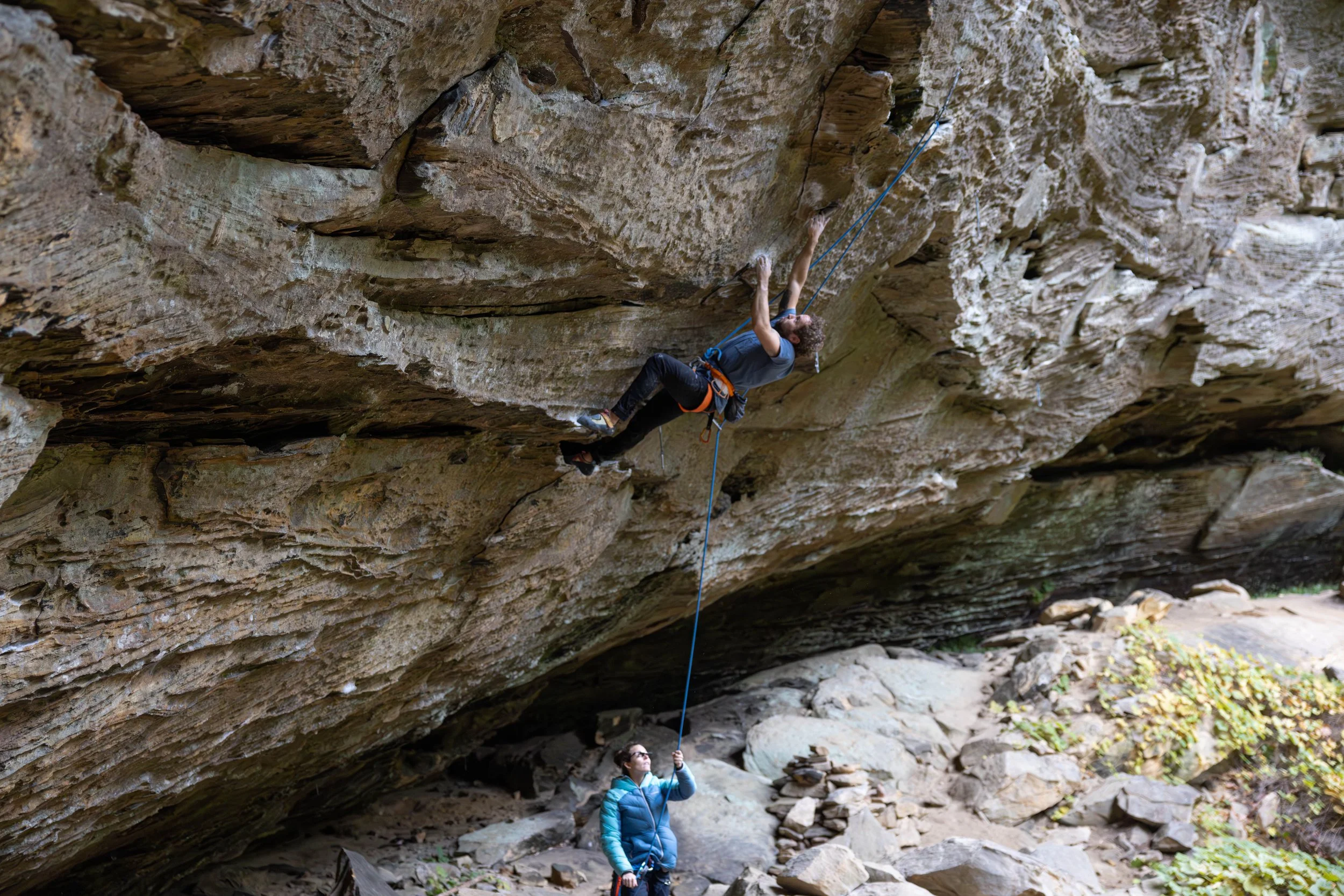 Rock climber ascending a steep rock face with a safety rope, belayer below holding the rope.