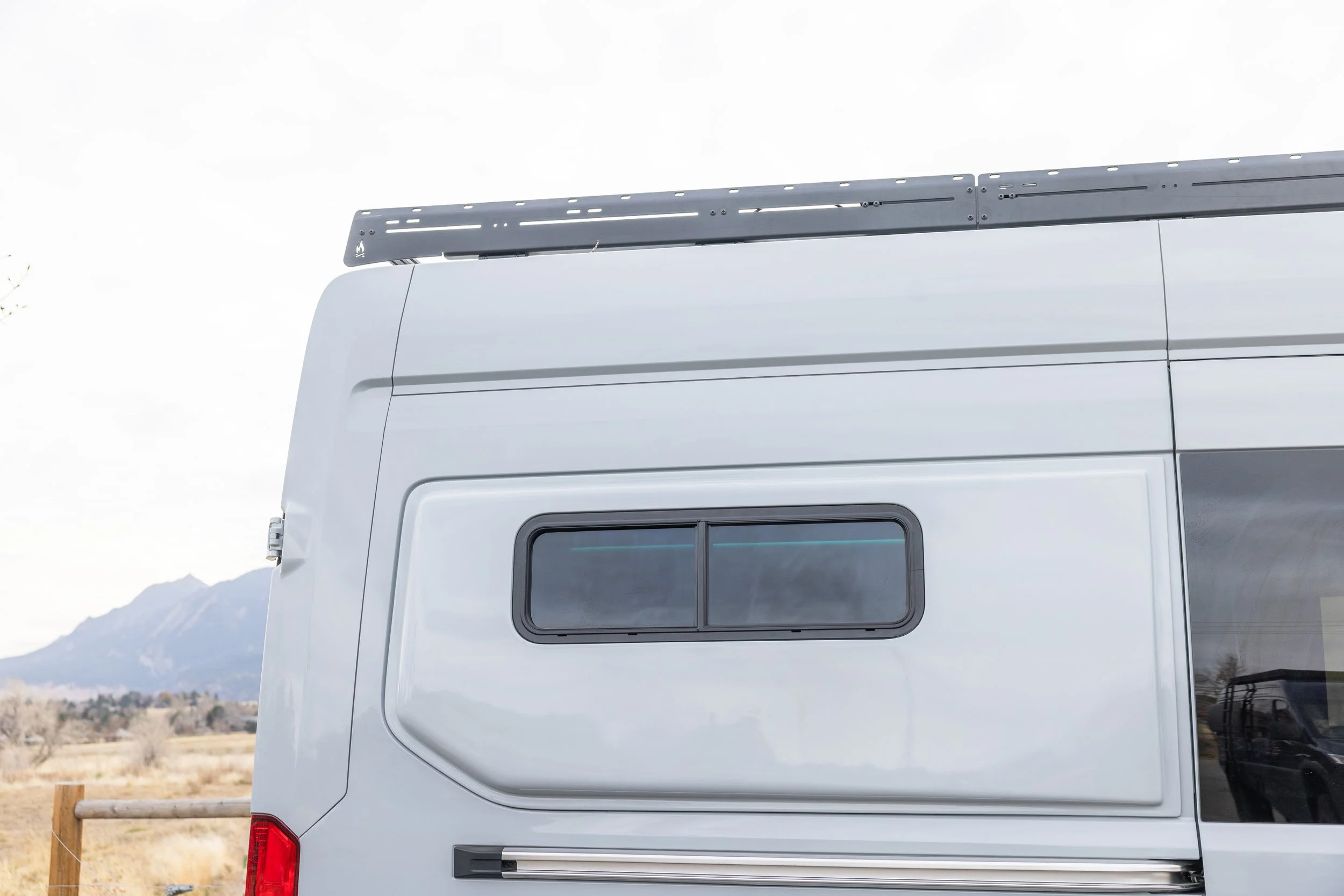 Close-up of the side of a white camper van with a window and a black roof rack, with a mountainous landscape in the background