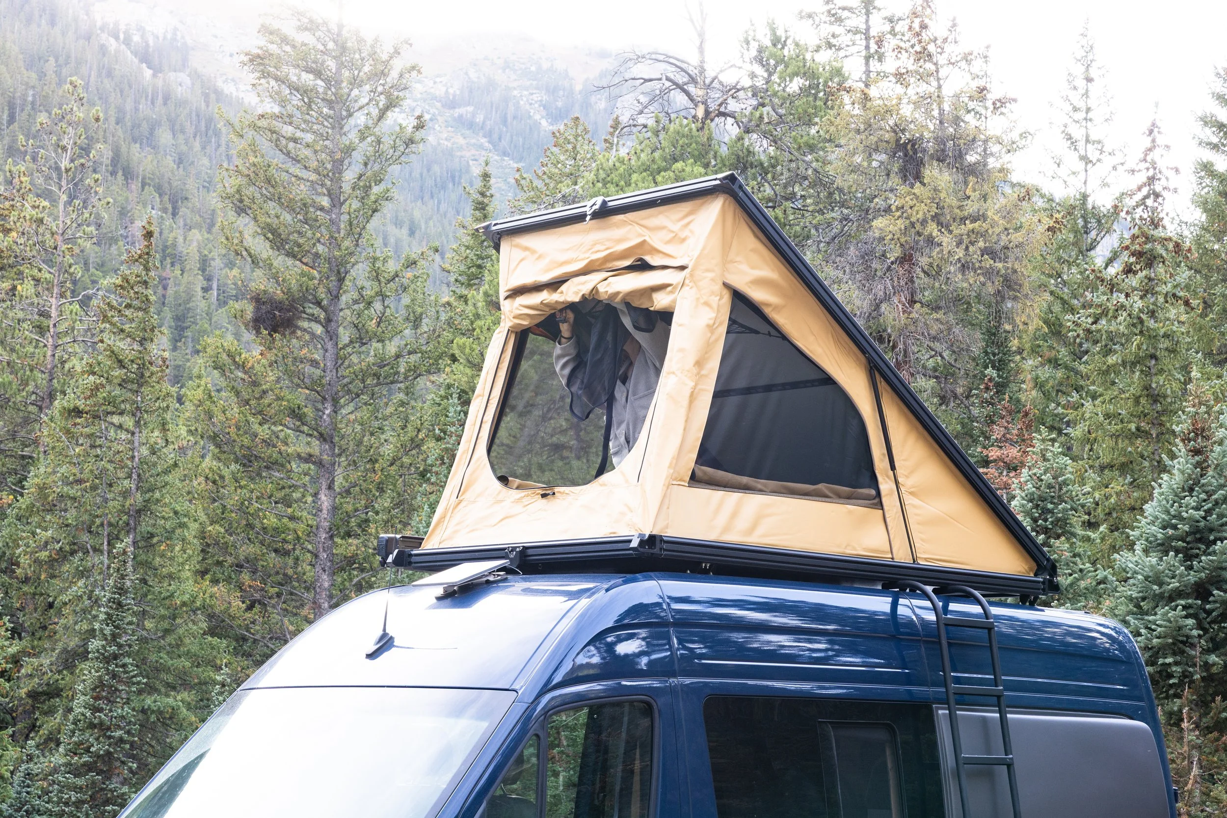 A blue camper van with a pop-up roof tent set up on top, parked in a forested area with trees and mountains in the background.