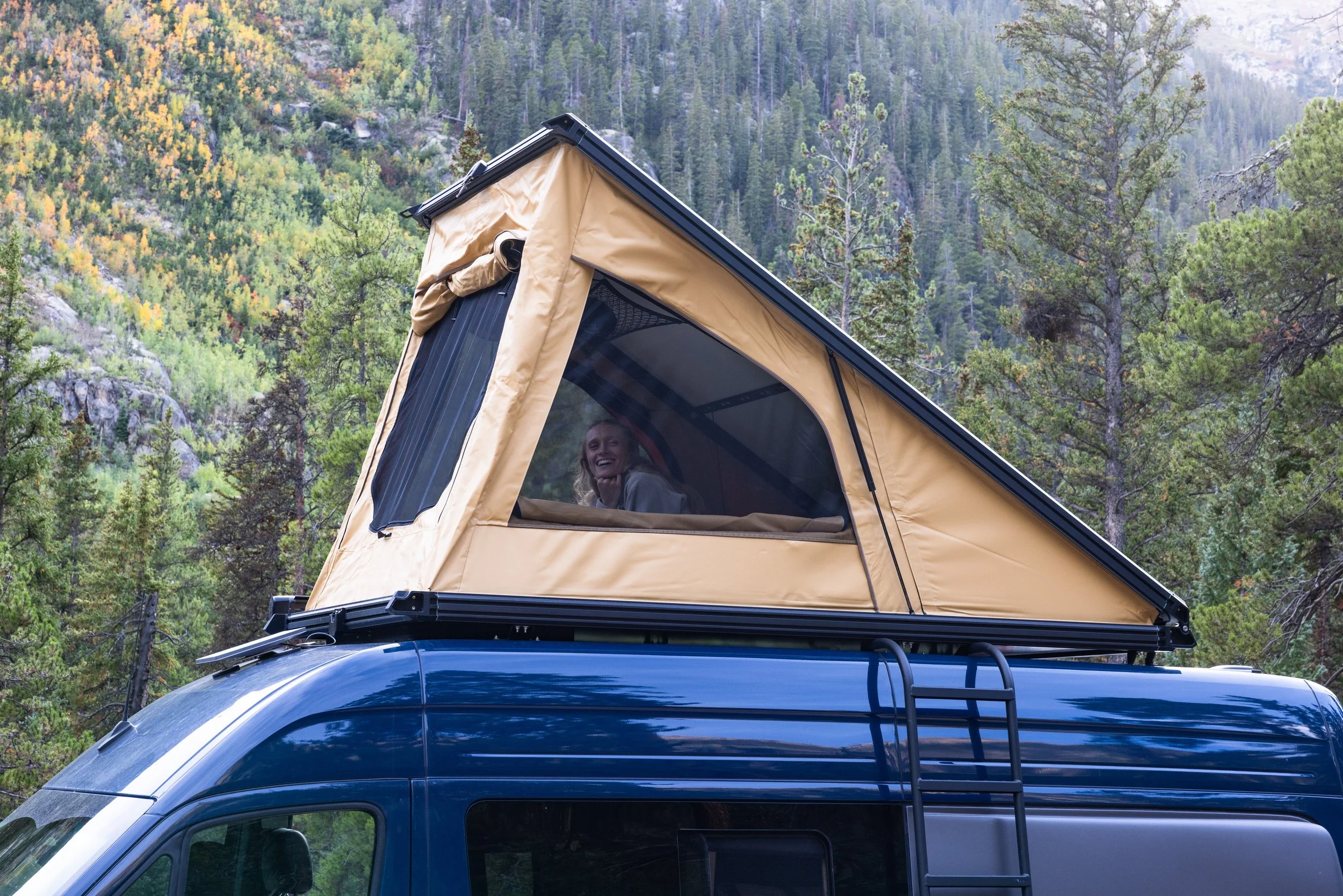 A woman smiling inside a small tent mounted on top of a blue camper van, set against a forested mountain landscape.