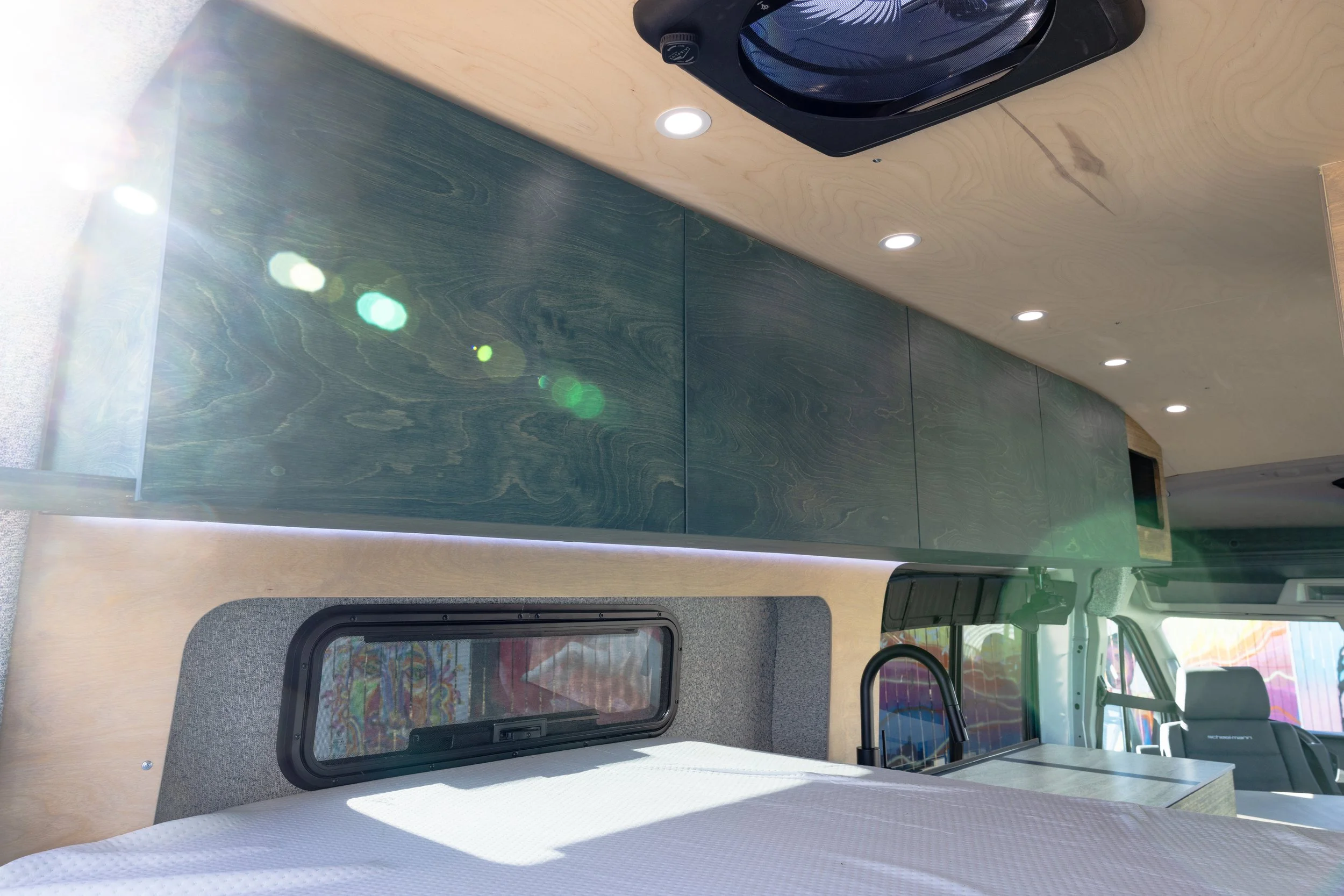 Interior of a camper van showing a wooden ceiling, cabinets with a blue-green wood finish, LED ceiling lights, and a small window with a curtain.