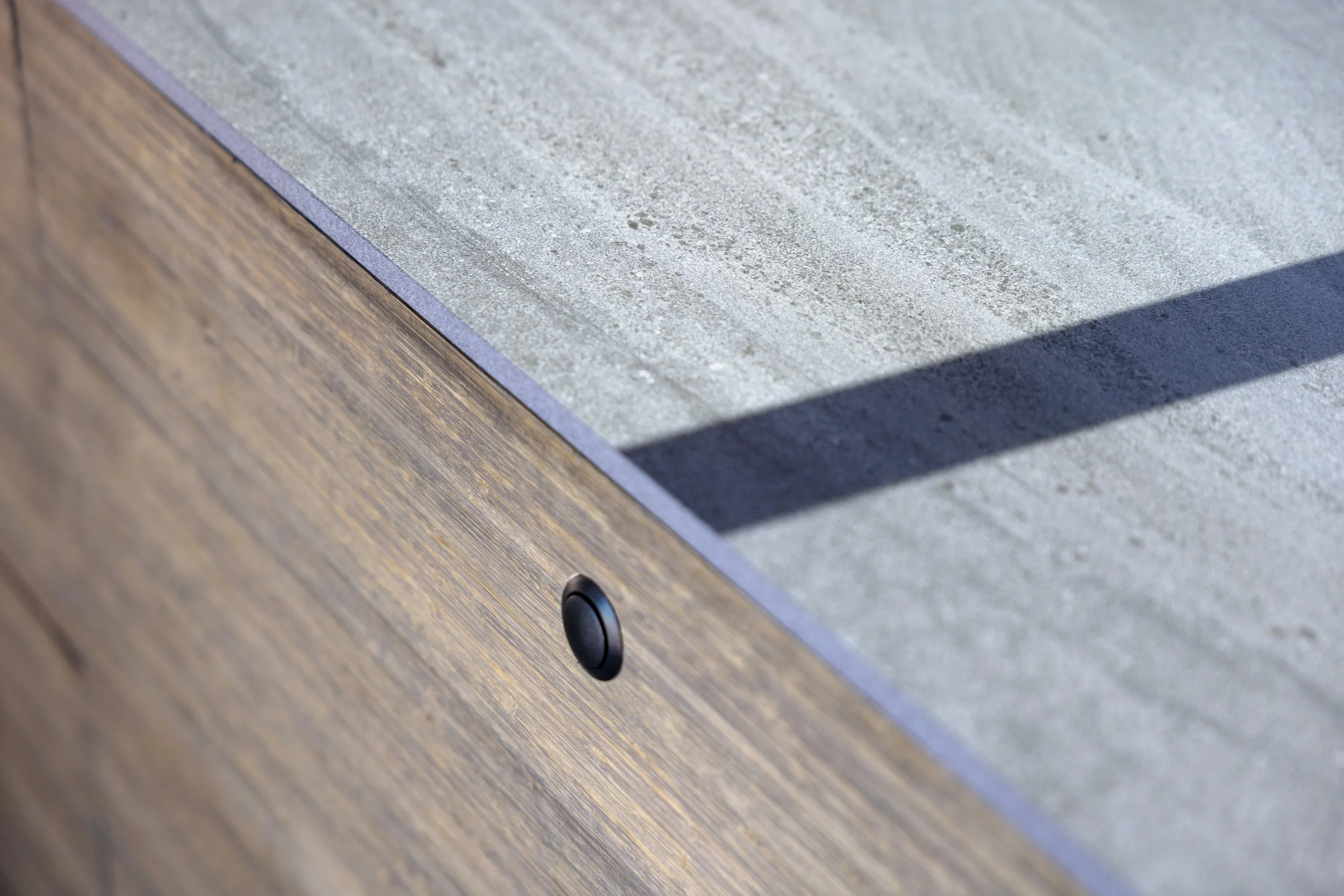 Close-up of a piece of furniture with a wood grain finish and a concrete top with a smooth surface, featuring a small black circular button or sensor on the wooden side.