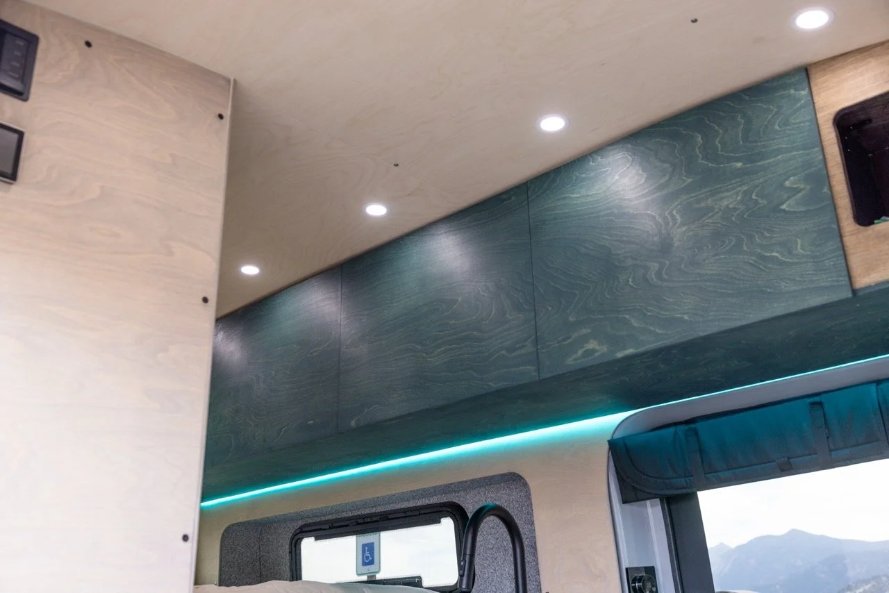 Interior of a modern camper van with beige and wood paneling, blue-green textured wall panels, ceiling lights, a window with a blue curtain, and a view of mountains outside.