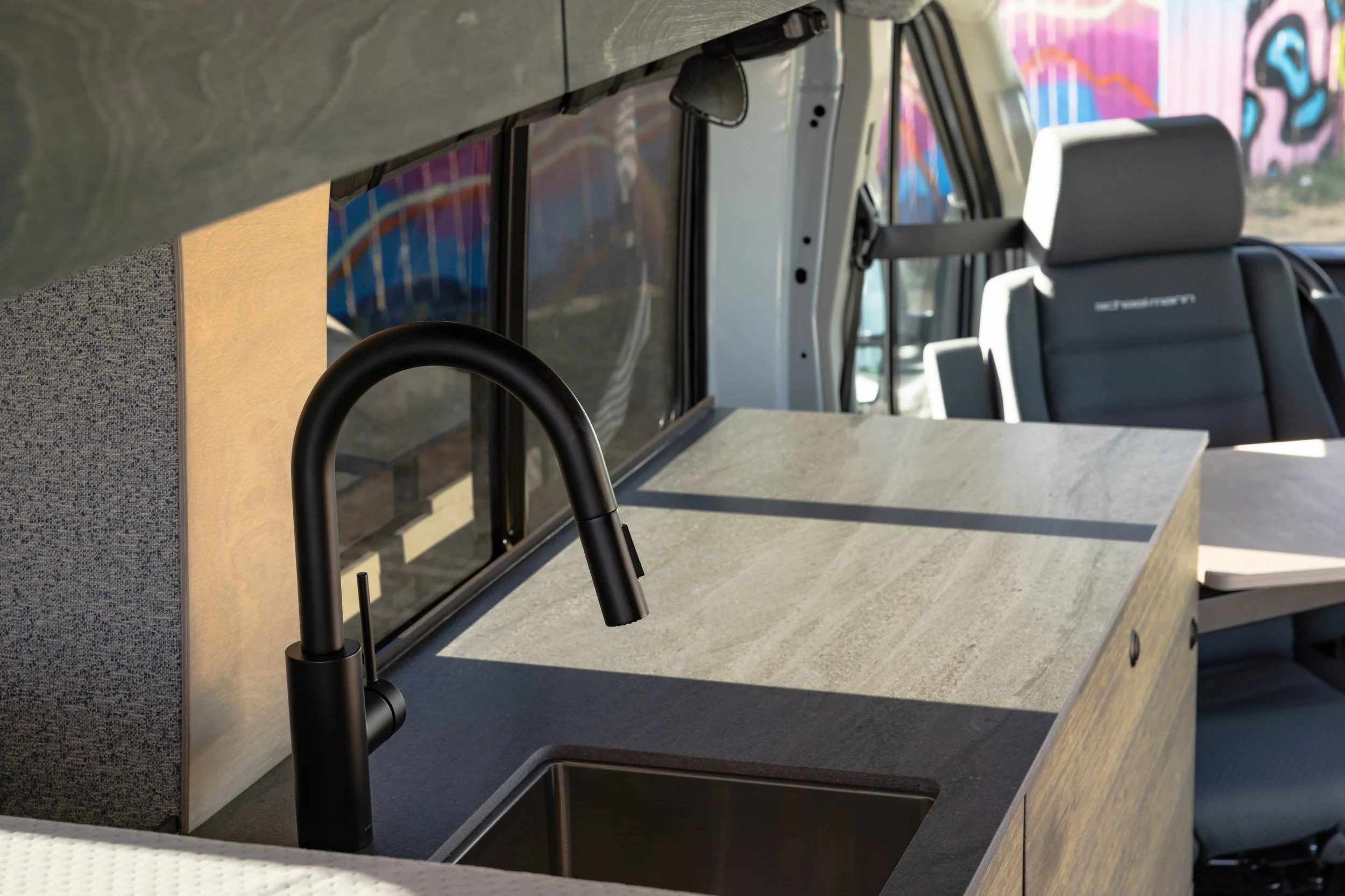 Interior of a van converted into a camper, showing a black faucet, a small sink, a countertop, and seating area with a gray driver's seat.