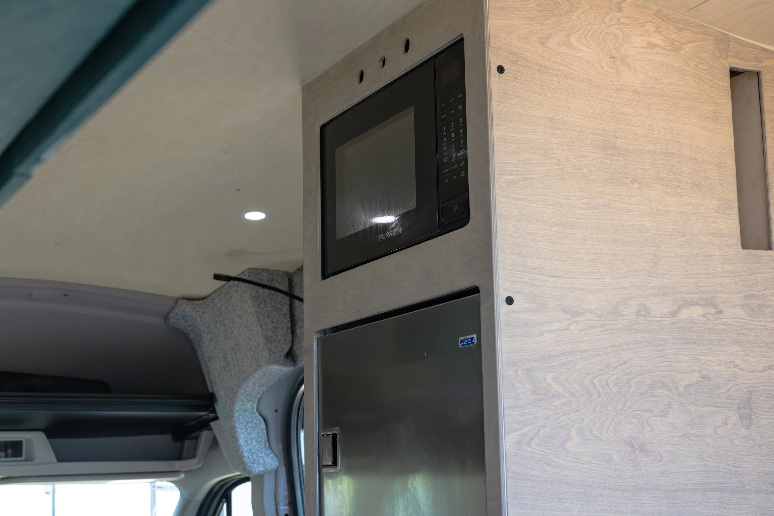 Interior of a camper with a microwave oven and a small refrigerator installed in a wooden cabinet.