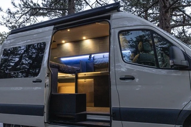 Interior of a custom camper van with a bed, lighting, and storage compartments, parked outdoors among trees.