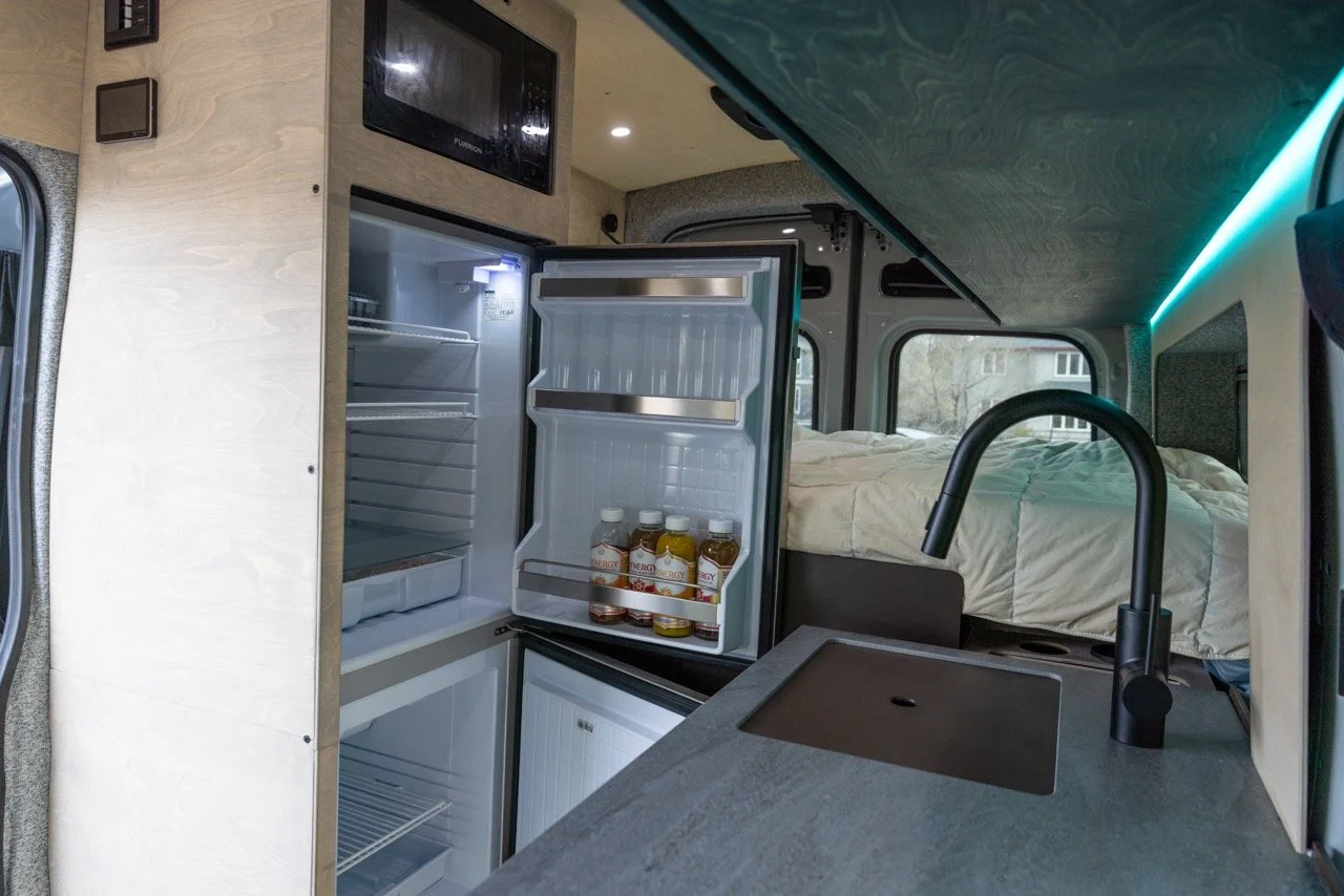 Open miniature refrigerator with three bottled drinks inside, in a compact living space with a bed, sink, and microwave.