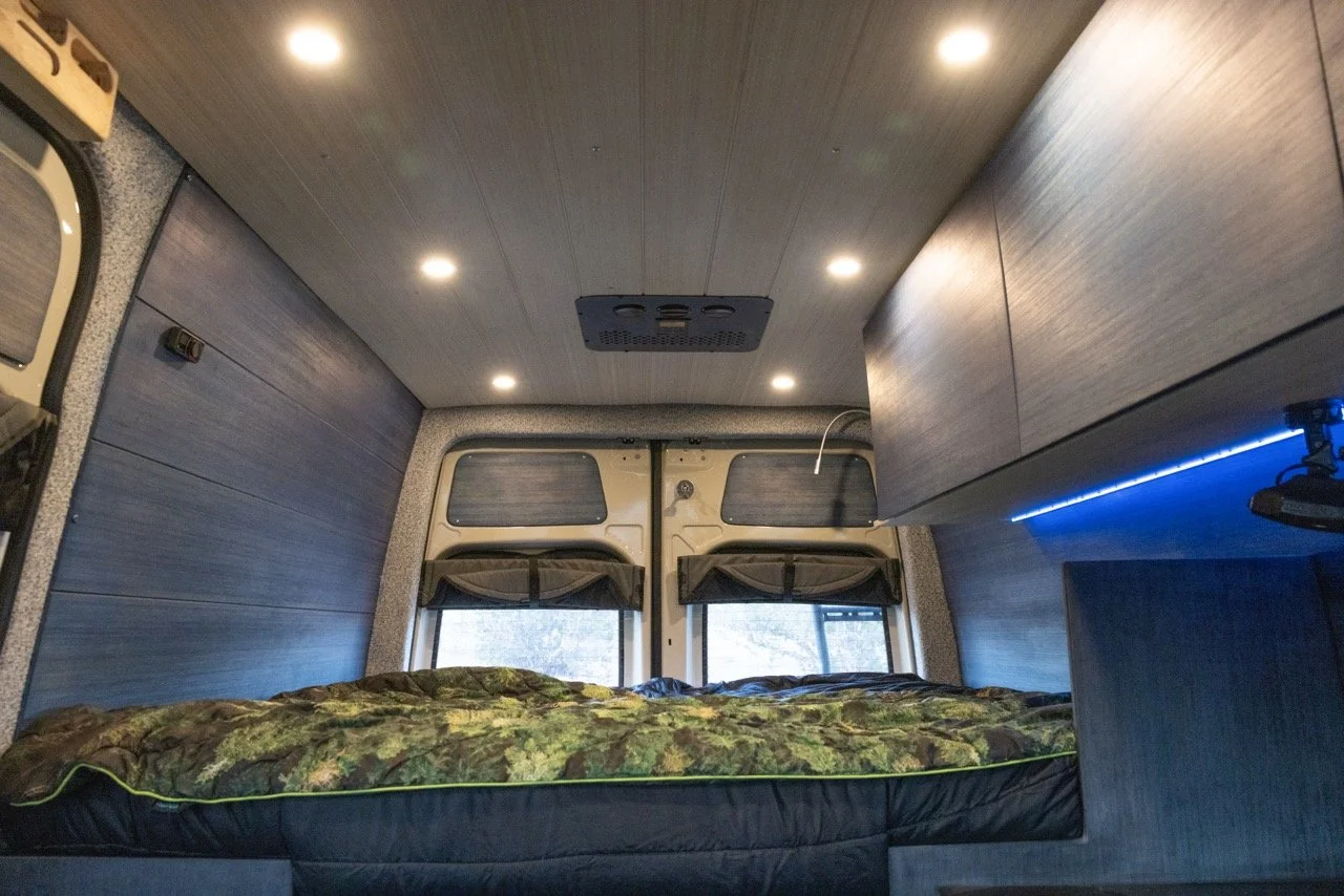 Interior of a camper van with a bed, overhead cabinets, LED strip lighting, and small windows with curtains.