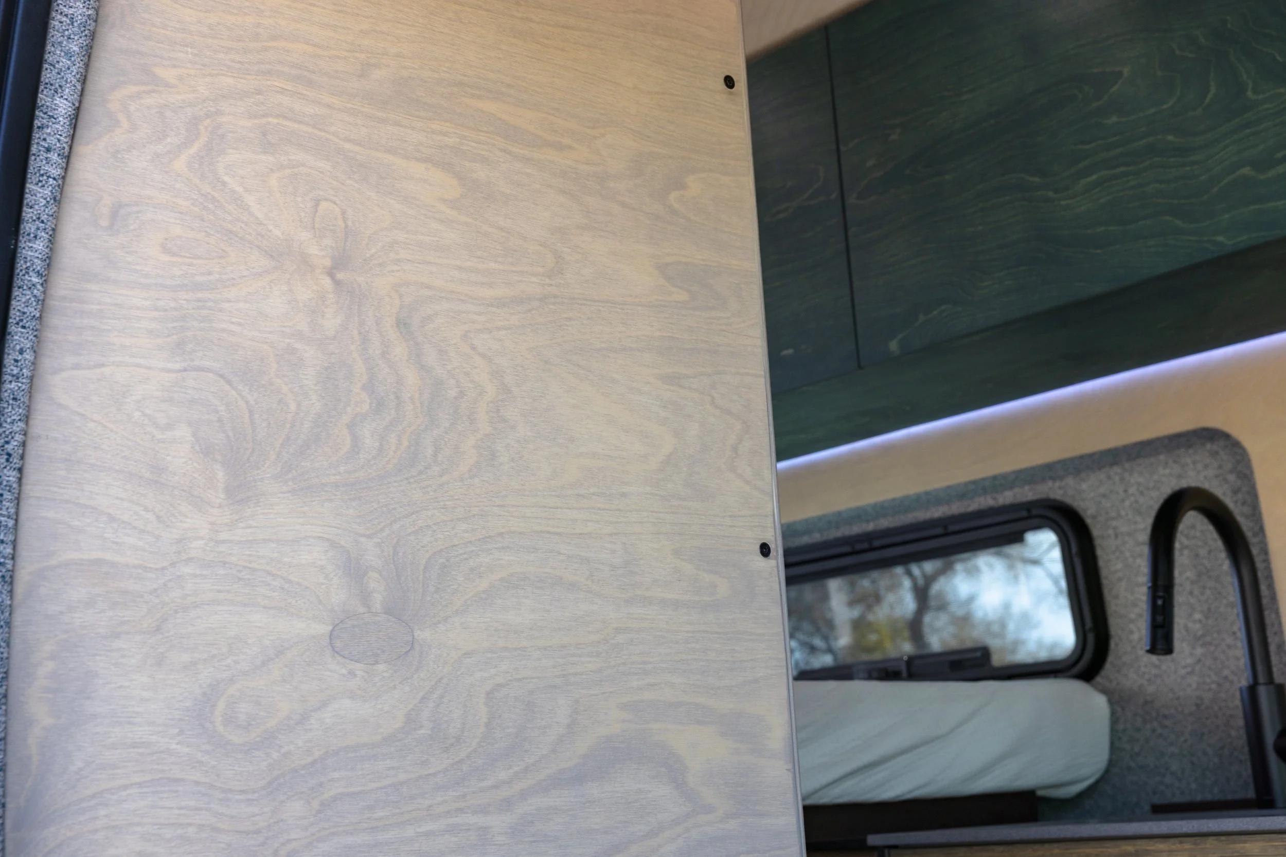 Interior of a camper van showing a light wood tabletop, a dark green cabinet, a window with trees outside, and a small bed with a rolled-up blanket.